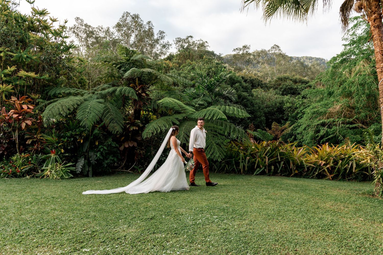 Bride and groom hold hands walking across green lawn with tropical rainforest backdrop.
