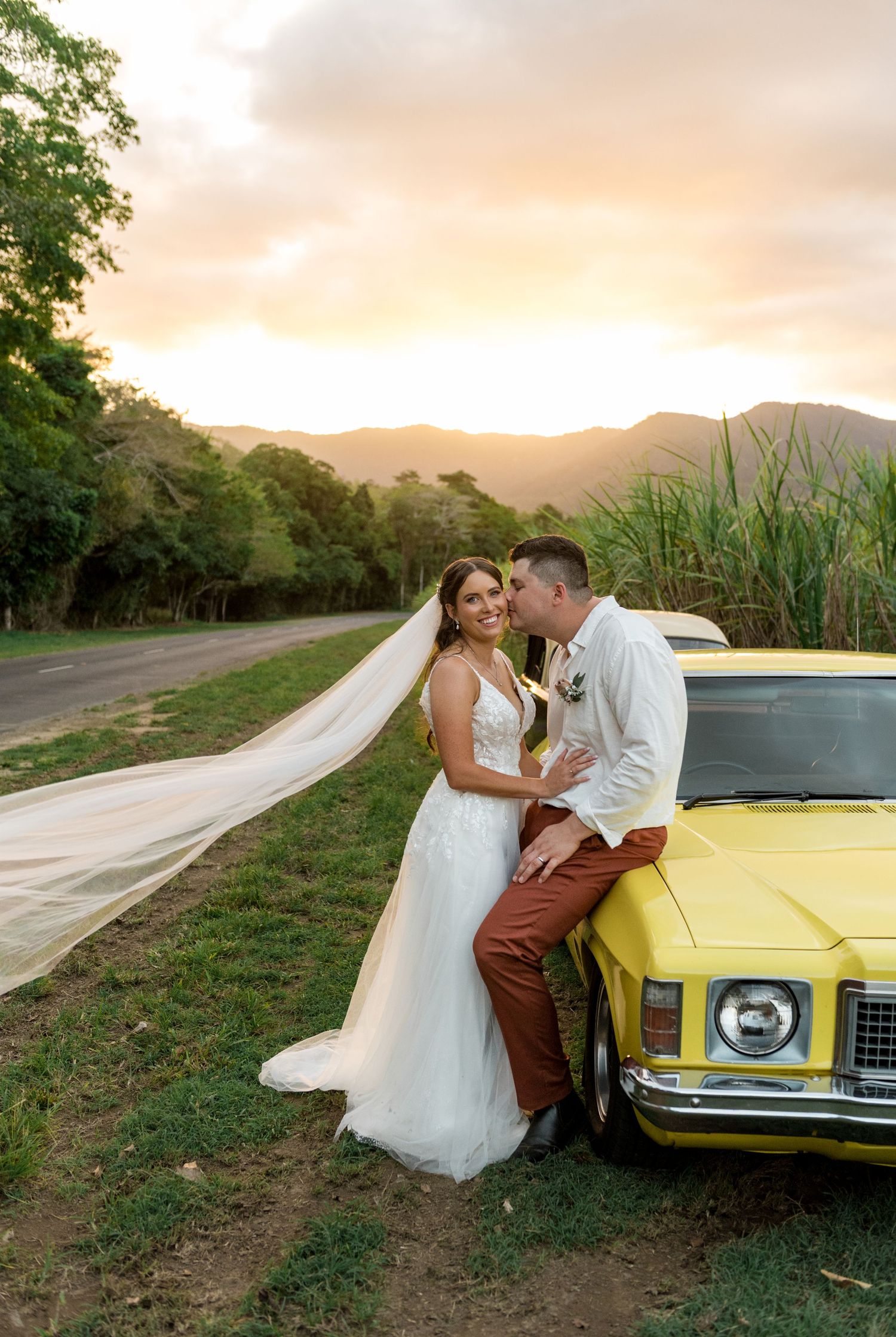 Groom kisses bride beside a vintage yellow car at a stunning golden sunset.