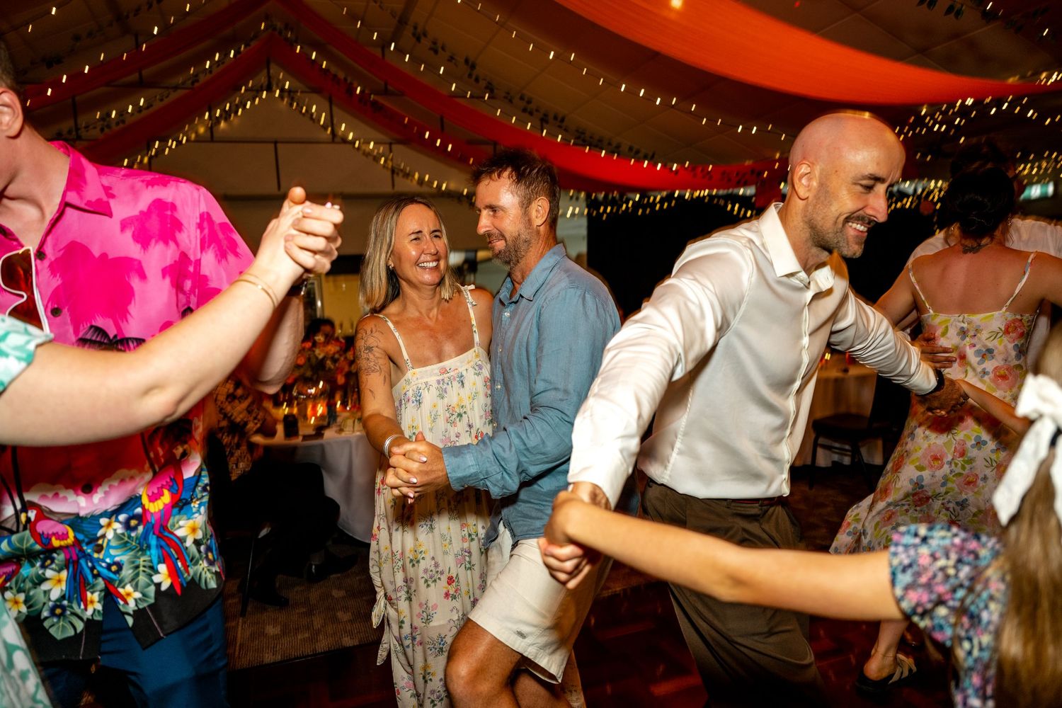 Guests dancing energetically at a wedding reception under fairy lights and red draping.
