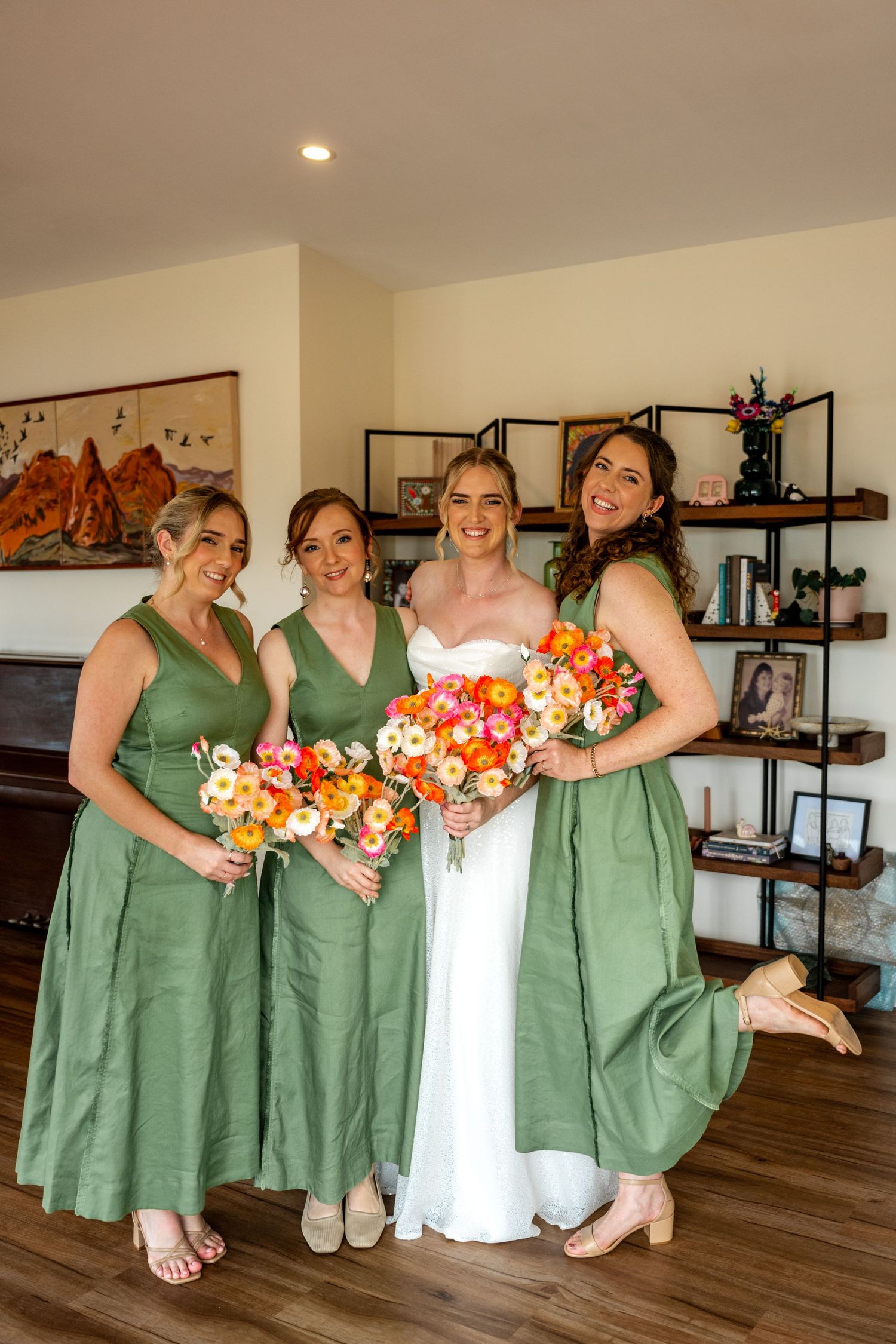 Bride with three bridesmaids in sage green dresses holding vibrant orange and pink bouquets indoors.
