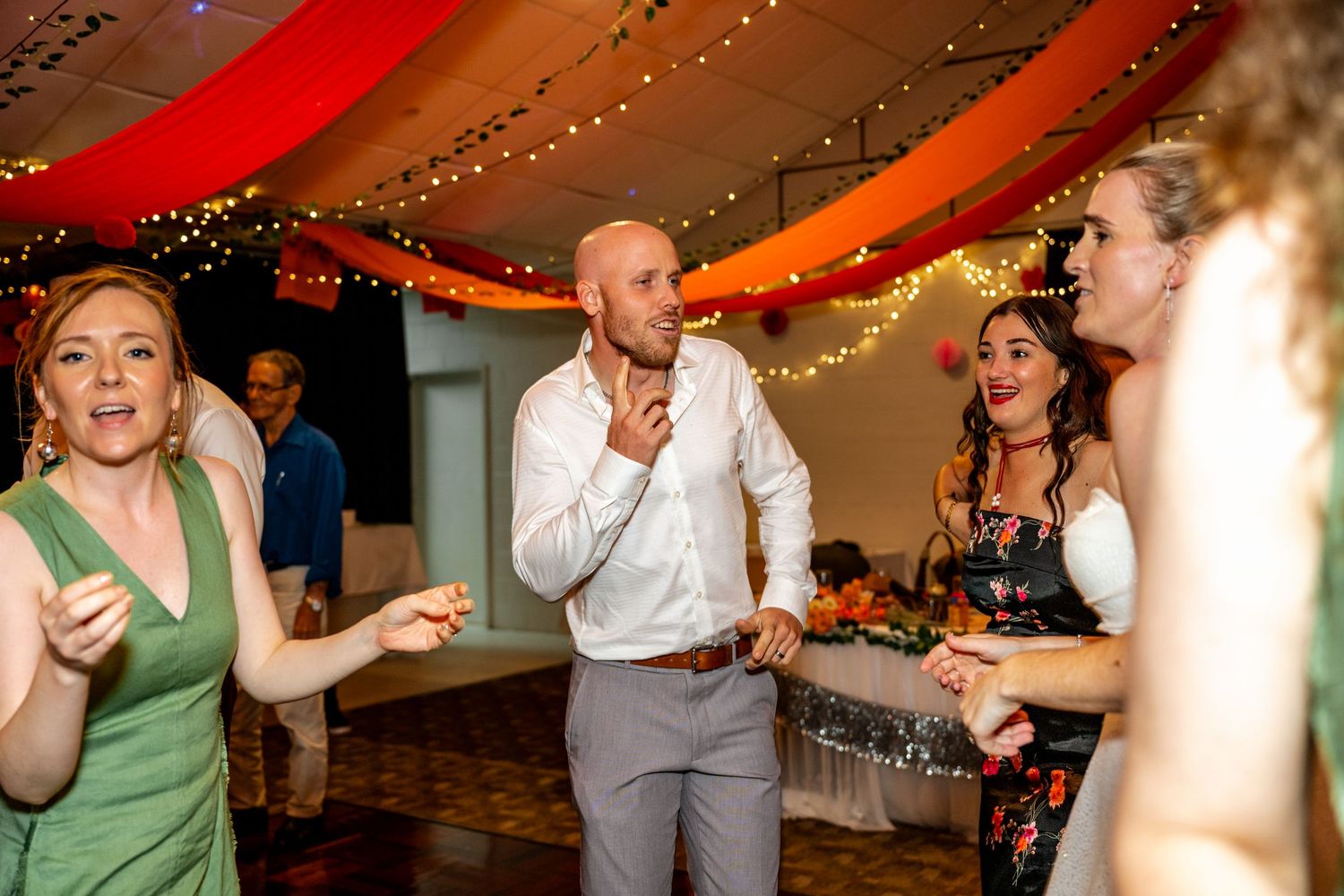 Wedding guests including man in white shirt dance together at a festive reception with fairy lights.