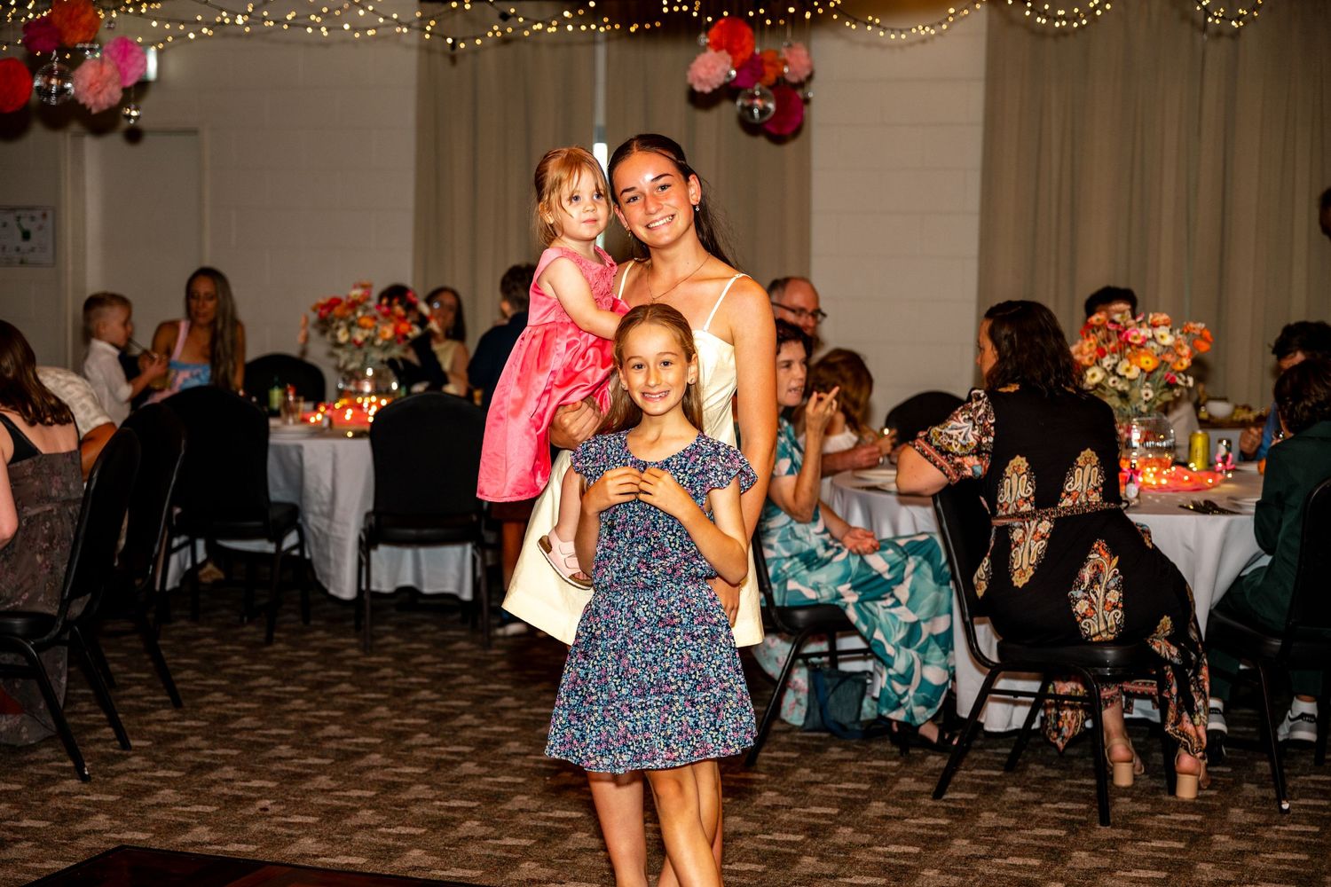 Woman in white dress posing with two young girls in colorful dresses at a festive wedding reception.