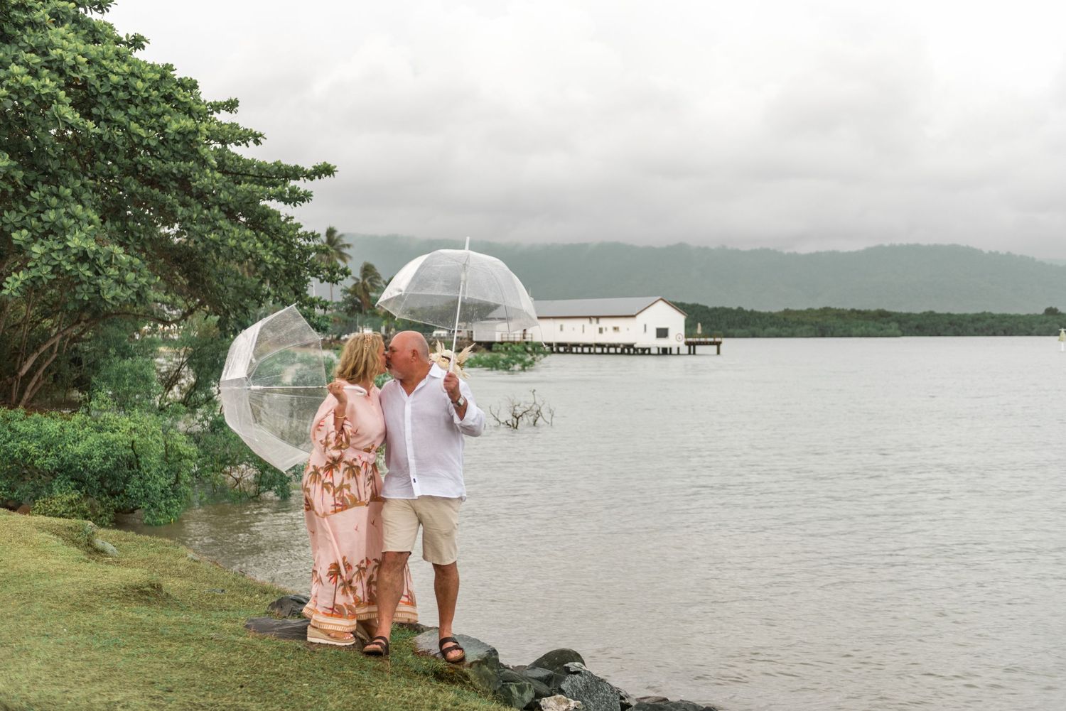 Couple standing by a rocky waterfront with a clear umbrella overlooking a tropical bay and pier.