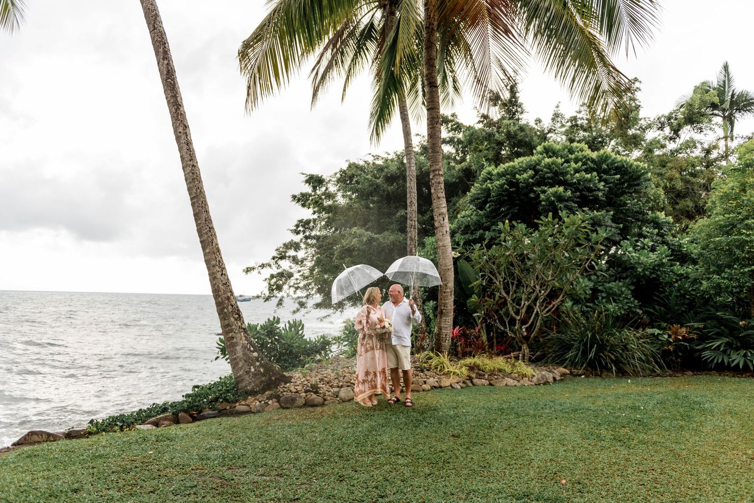 Mature couple holding clear umbrellas near a tropical oceanside garden with leaning palm trees.