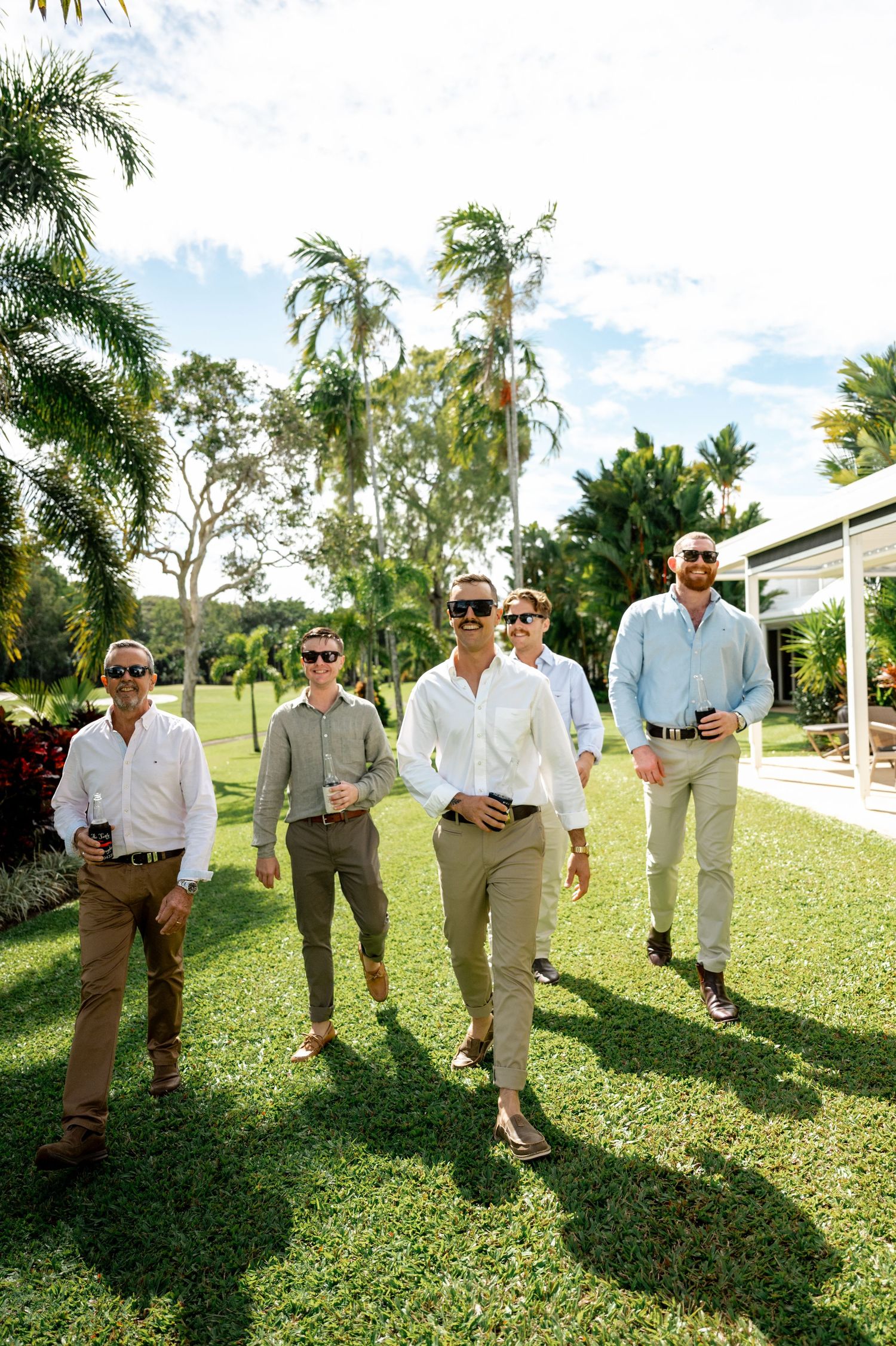 Five groomsmen in white and light blue shirts walking through tropical garden setting on wedding day.