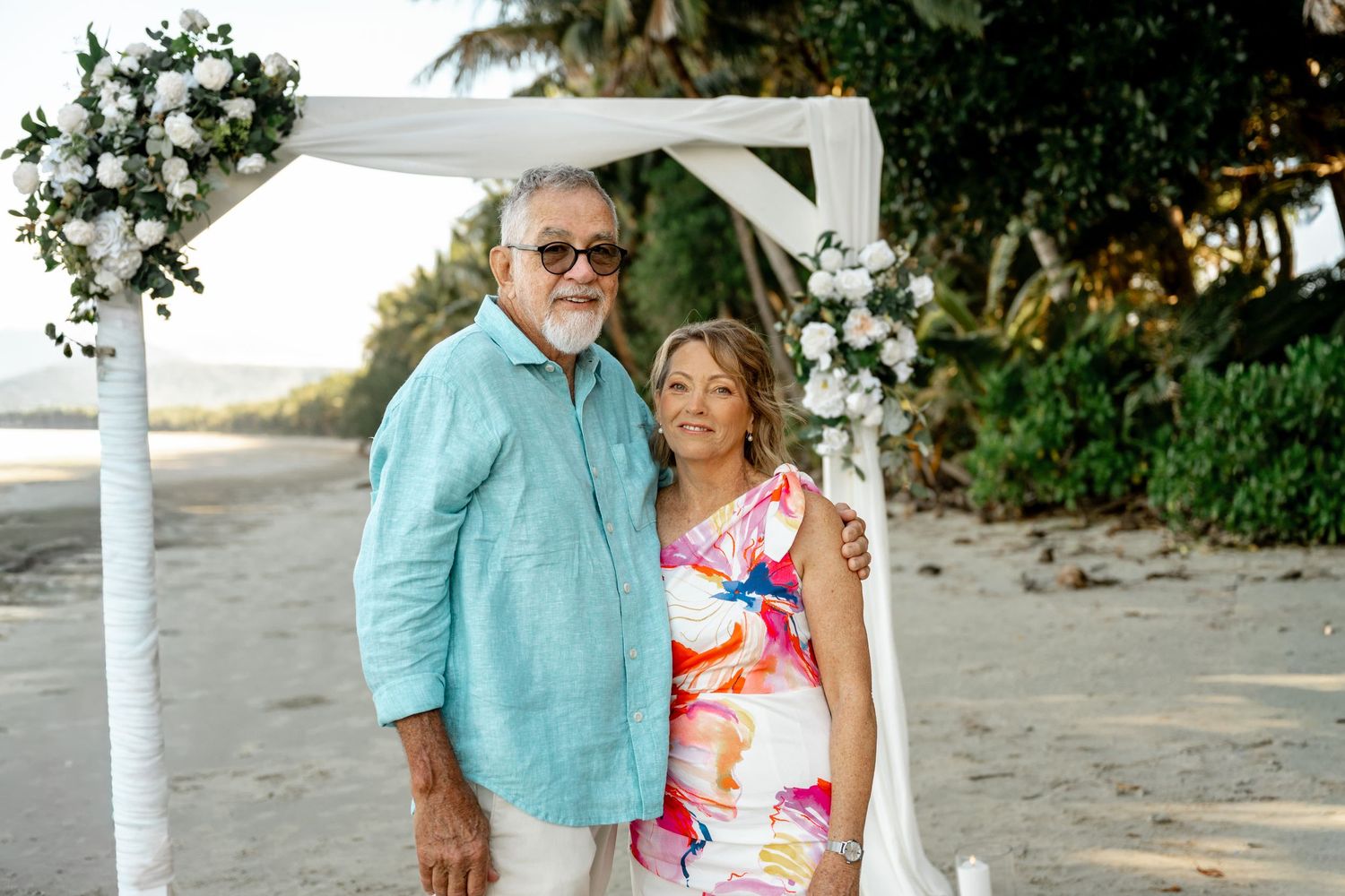 Older couple smiling together under a white floral wedding arch on a sandy tropical beach at Four Mile Beach, Port Douglas.