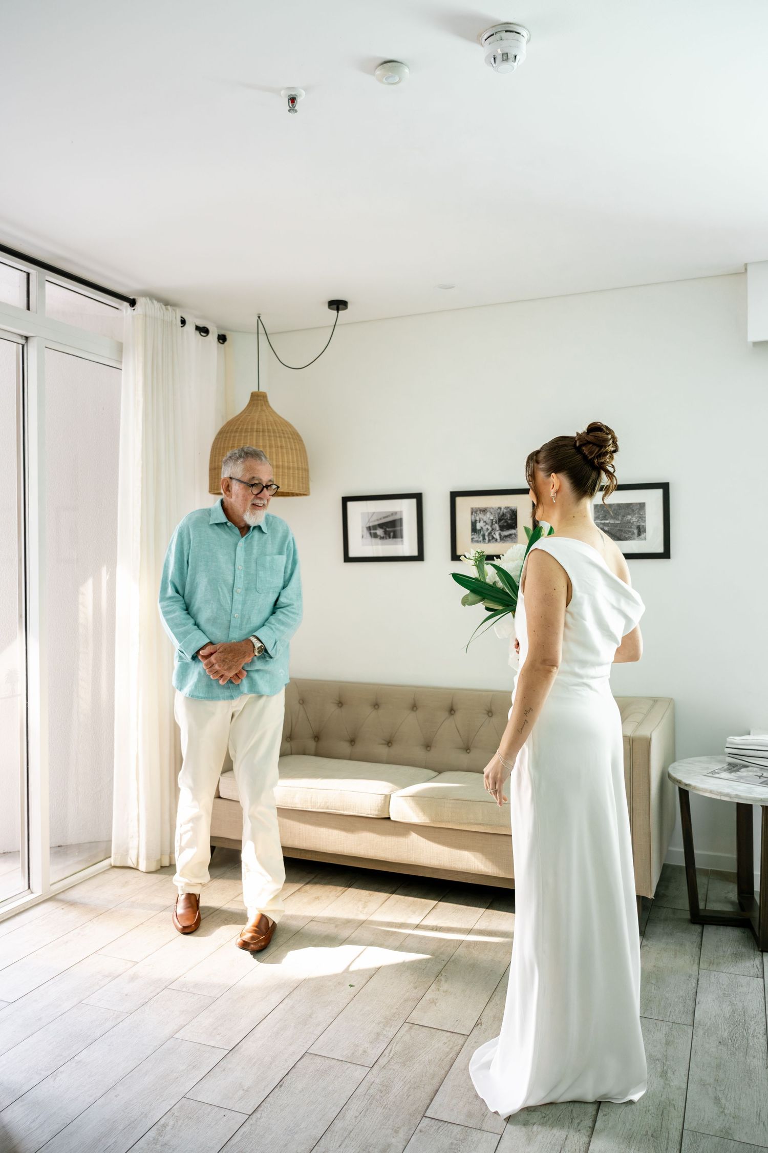 Older gentleman in turquoise shirt smiling at bride in white asymmetric wedding dress in bright modern room at Sheraton Grand Mirage Resort.
