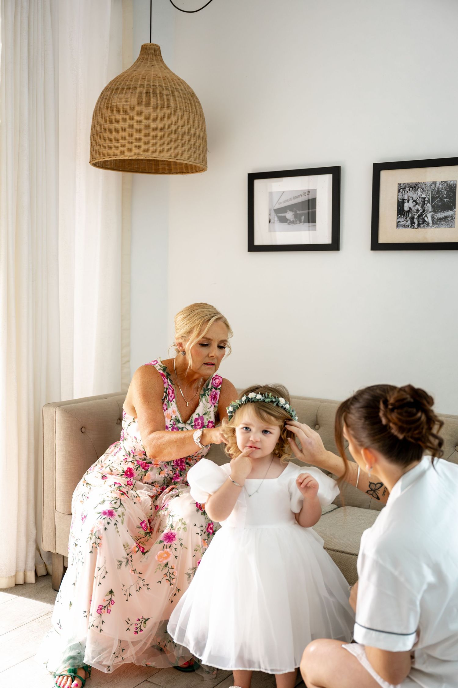 Woman in floral dress adjusts flower crown on little girl in white dress while bridesmaid assists indoors.