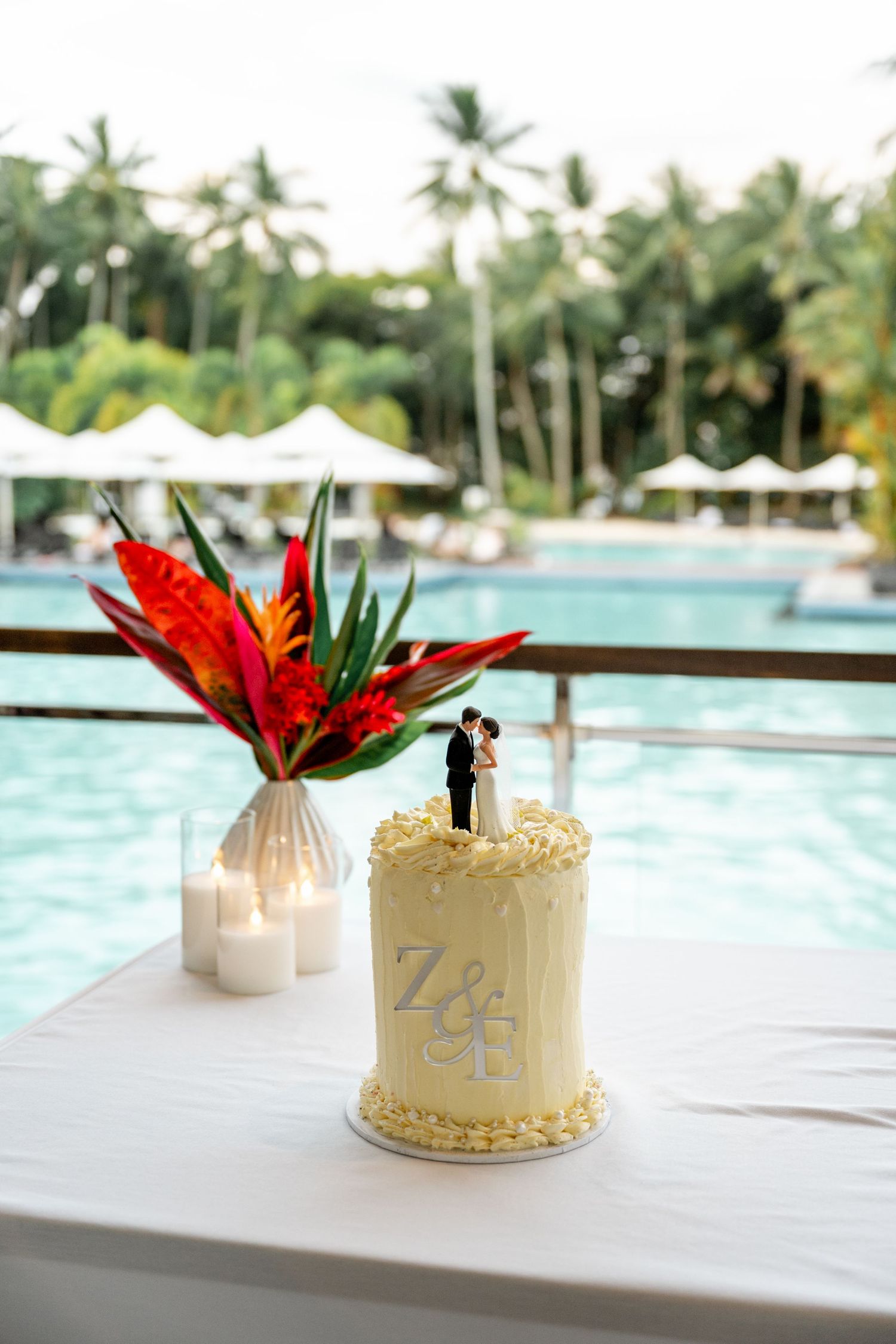 Elegant white wedding cake with Z&E monogram topper beside tropical flowers near a resort pool at Sheraton Grand Mirage Port Douglas.