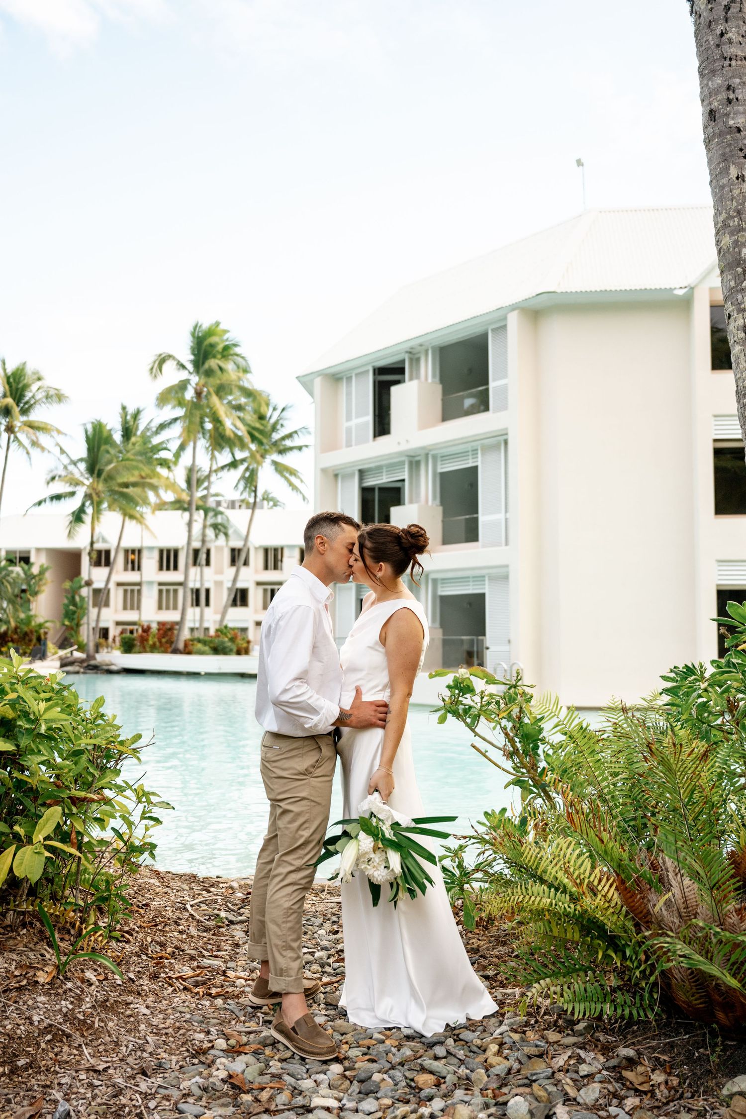 Bride and groom sharing a romantic kiss beside a tropical resort pool surrounded by palm trees at Sheraton Grand Mirage Port Douglas.