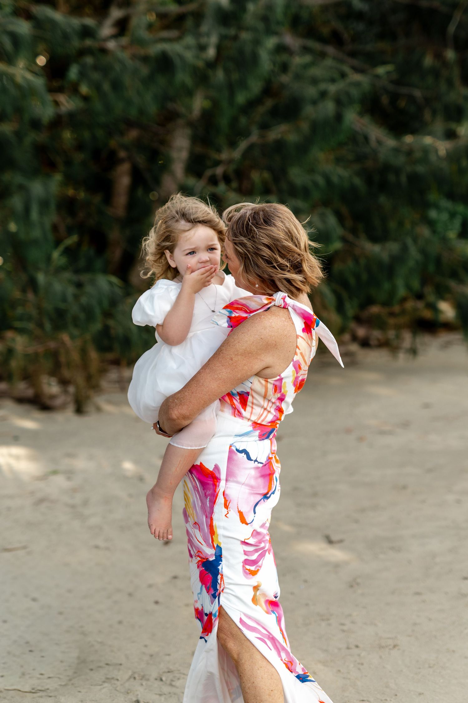 Woman in colorful floral dress carrying a young girl in white on a sandy beach surrounded by trees.