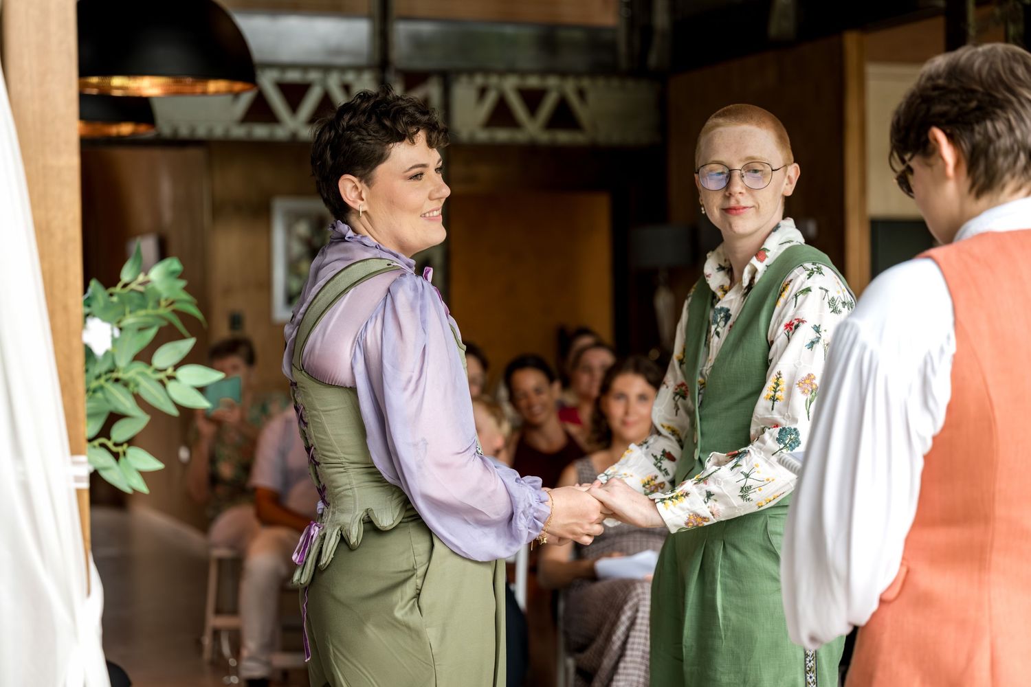 Two brides in green suits exchanging vows and holding hands at wedding altar with officiant.