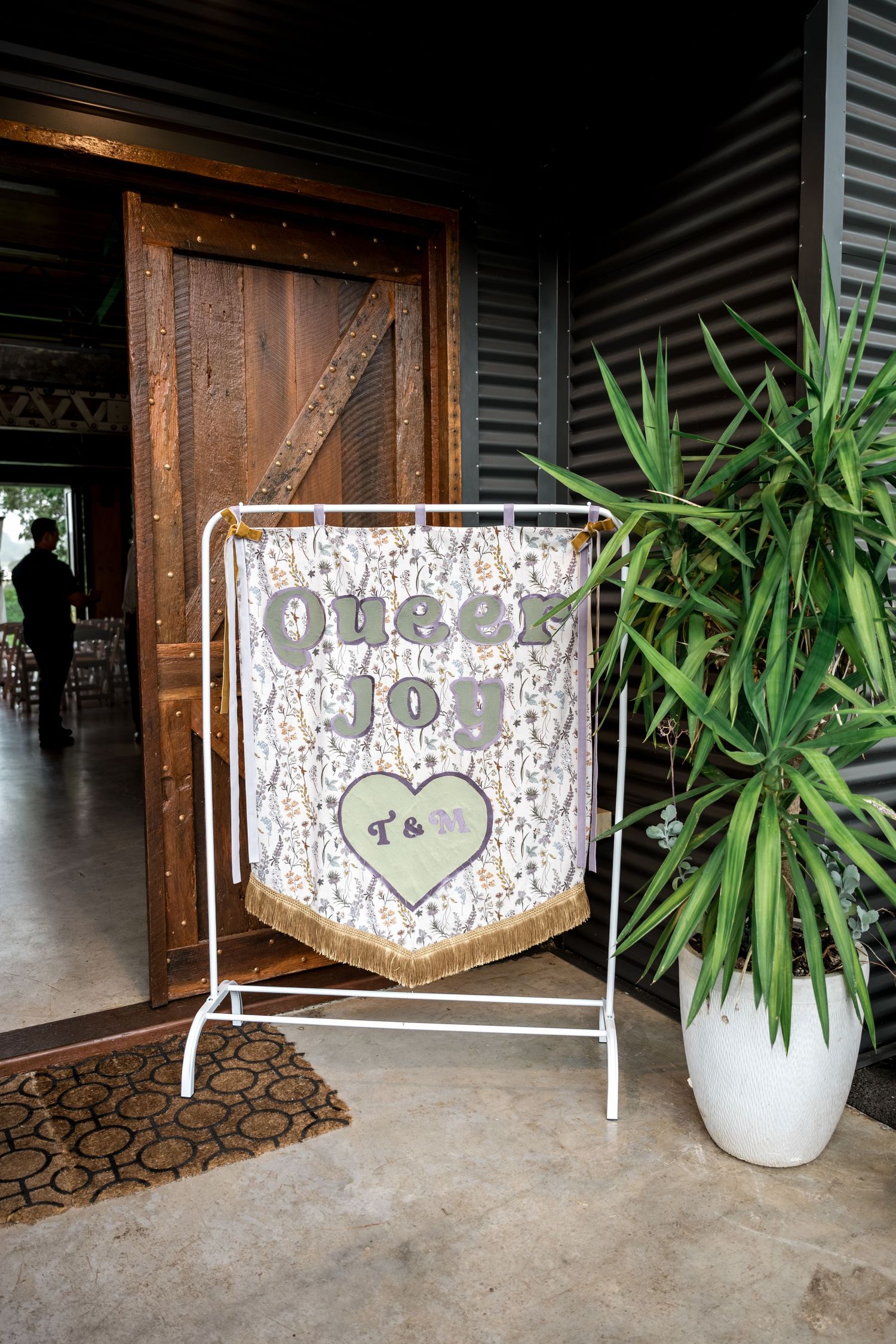 Queer Joy wedding banner with T&M heart displayed on white stand near rustic barn entrance.