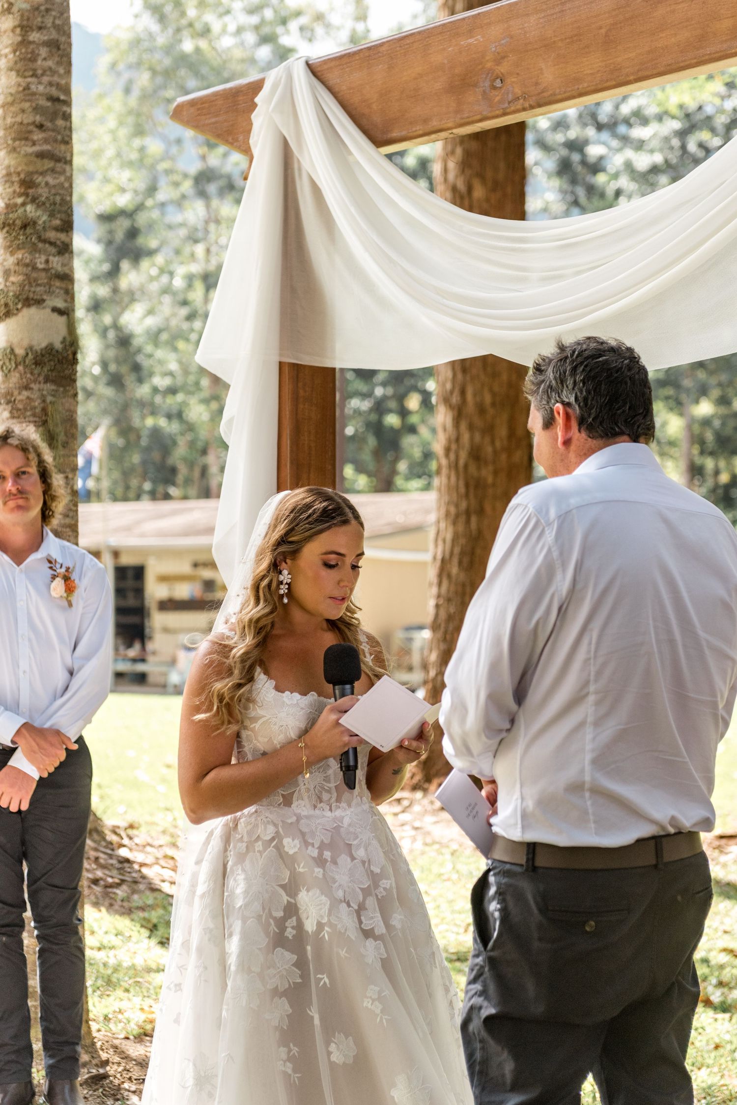 Bride in floral lace gown reads vows into microphone at outdoor woodland wedding ceremony.
