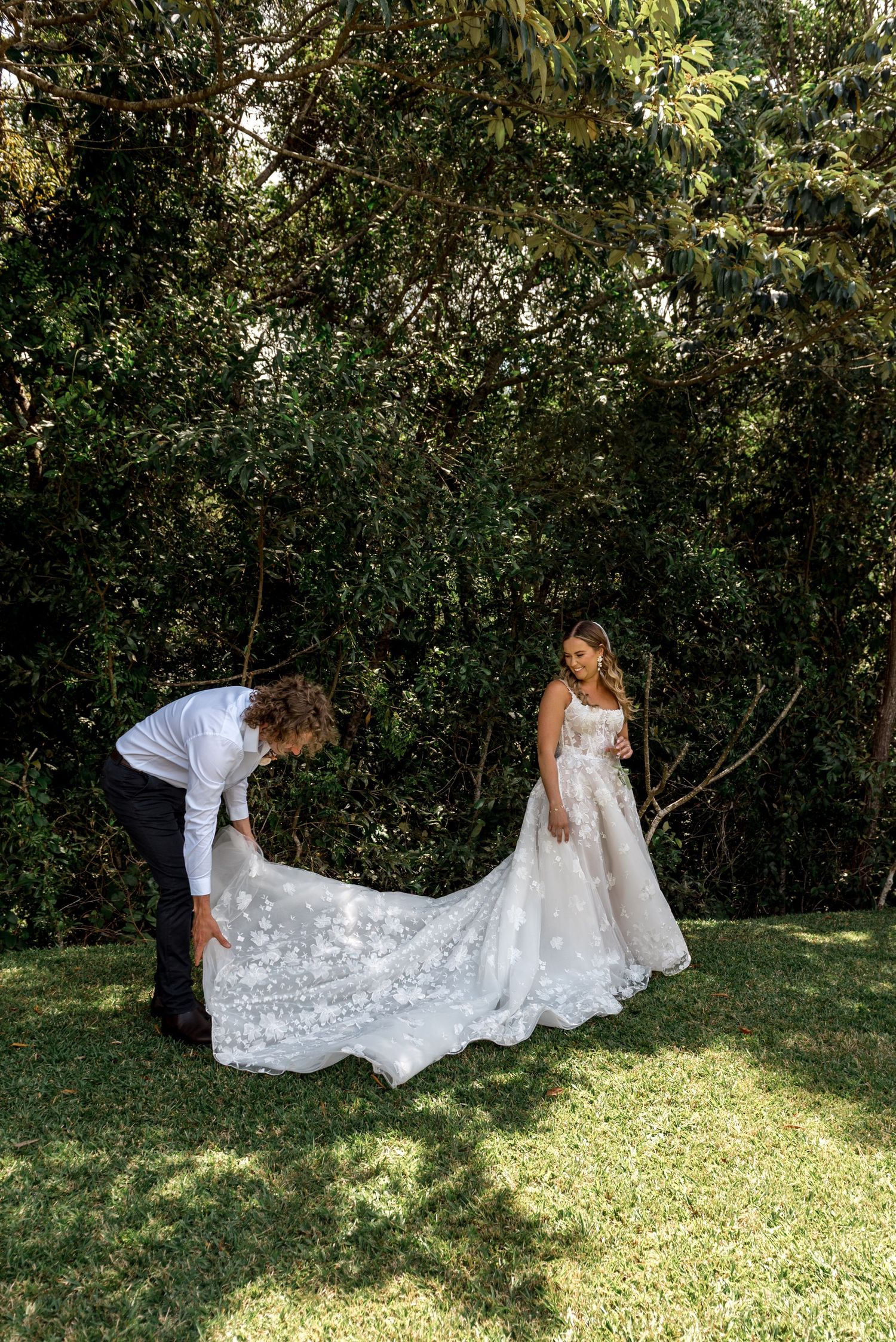 Groom carefully arranges long floral lace bridal train on grass before lush green trees.