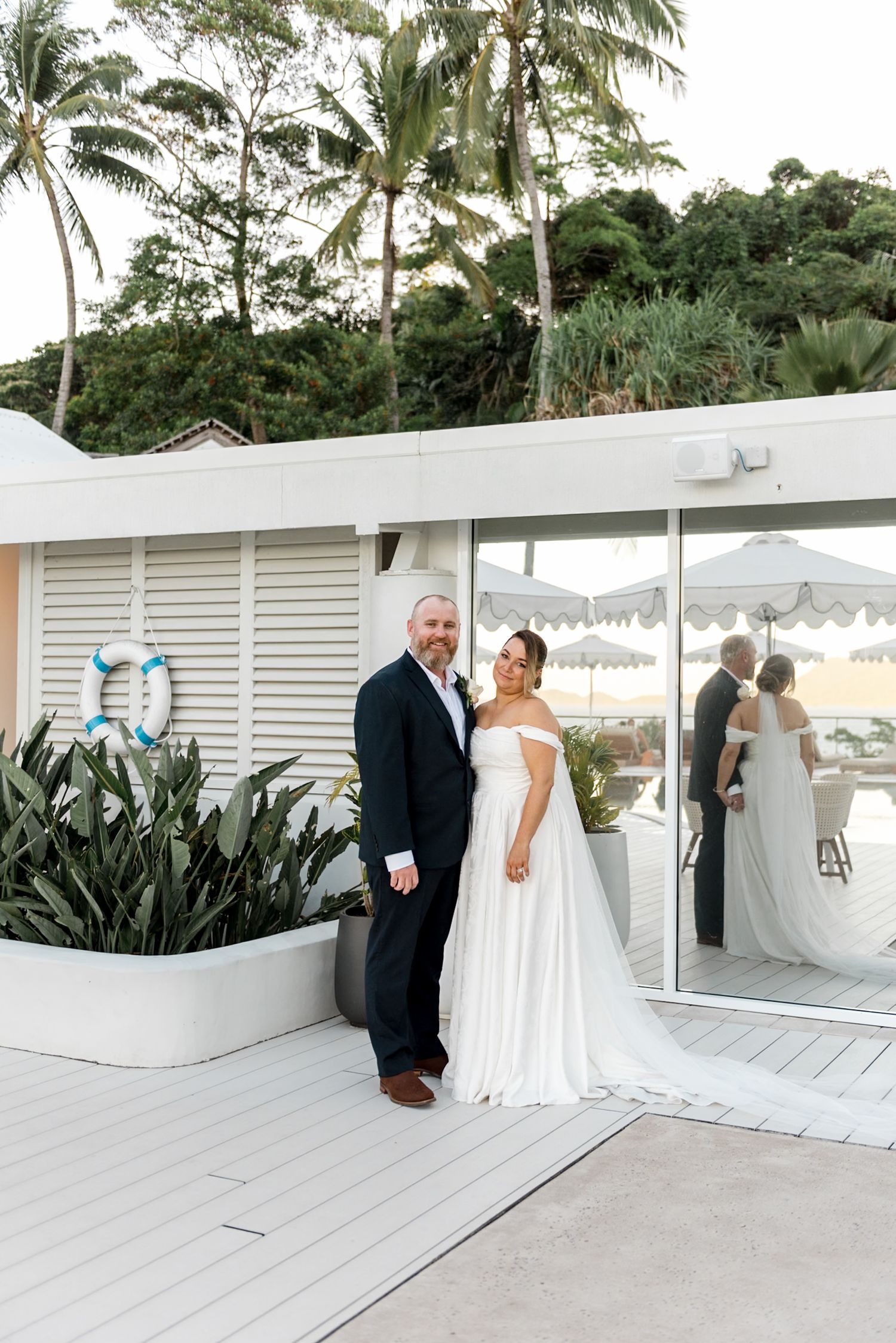Bride and groom posing together on a white beachside deck surrounded by palm trees.