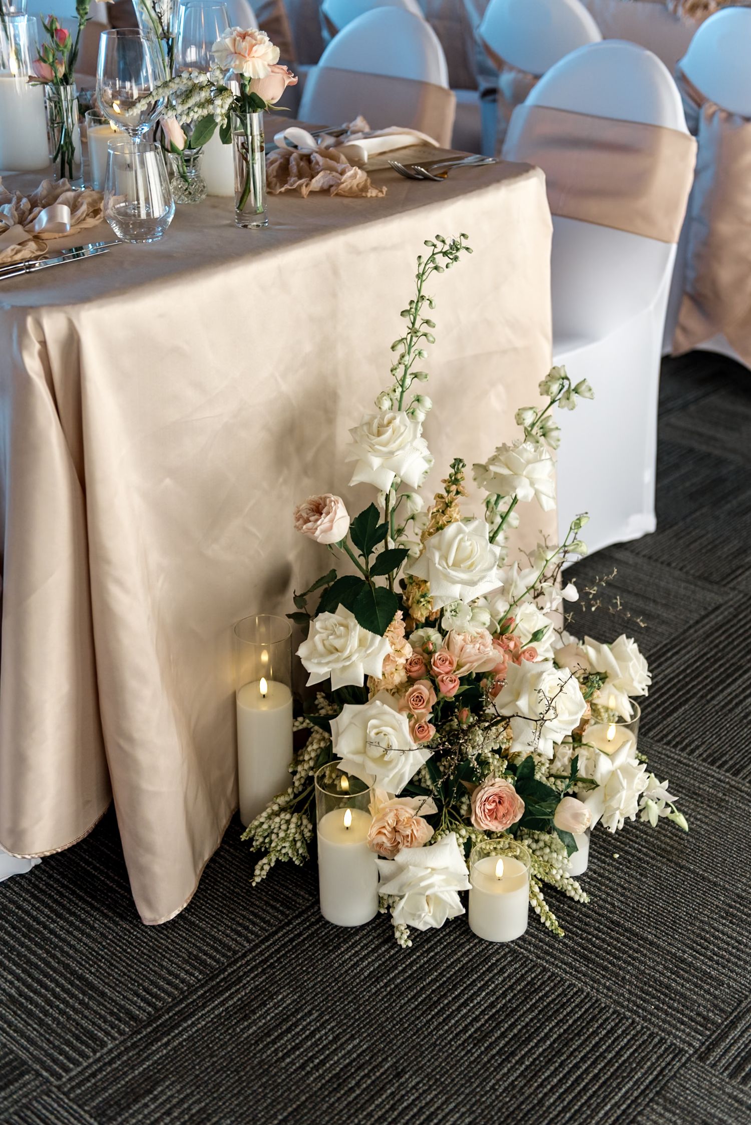 Floor floral arrangement of white and blush roses with pillar candles beside an elegant wedding reception table.