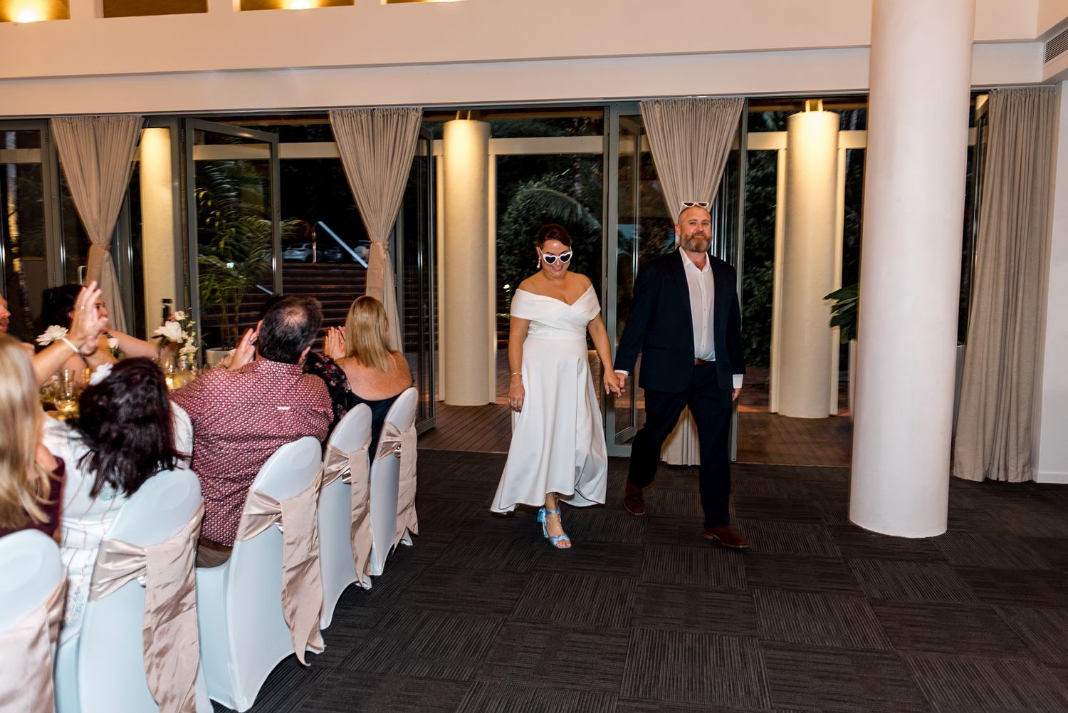 Newlyweds walking hand in hand through elegant draped reception hall as seated guests look on.