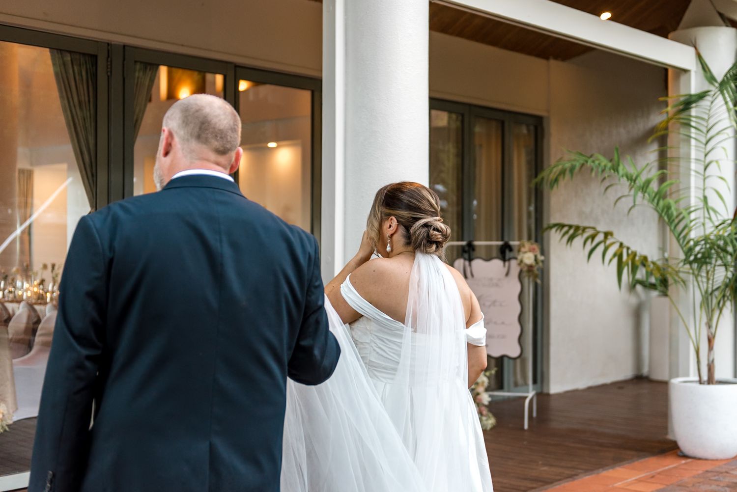 Bride and groom walking toward elegant venue entrance with veil flowing behind her.