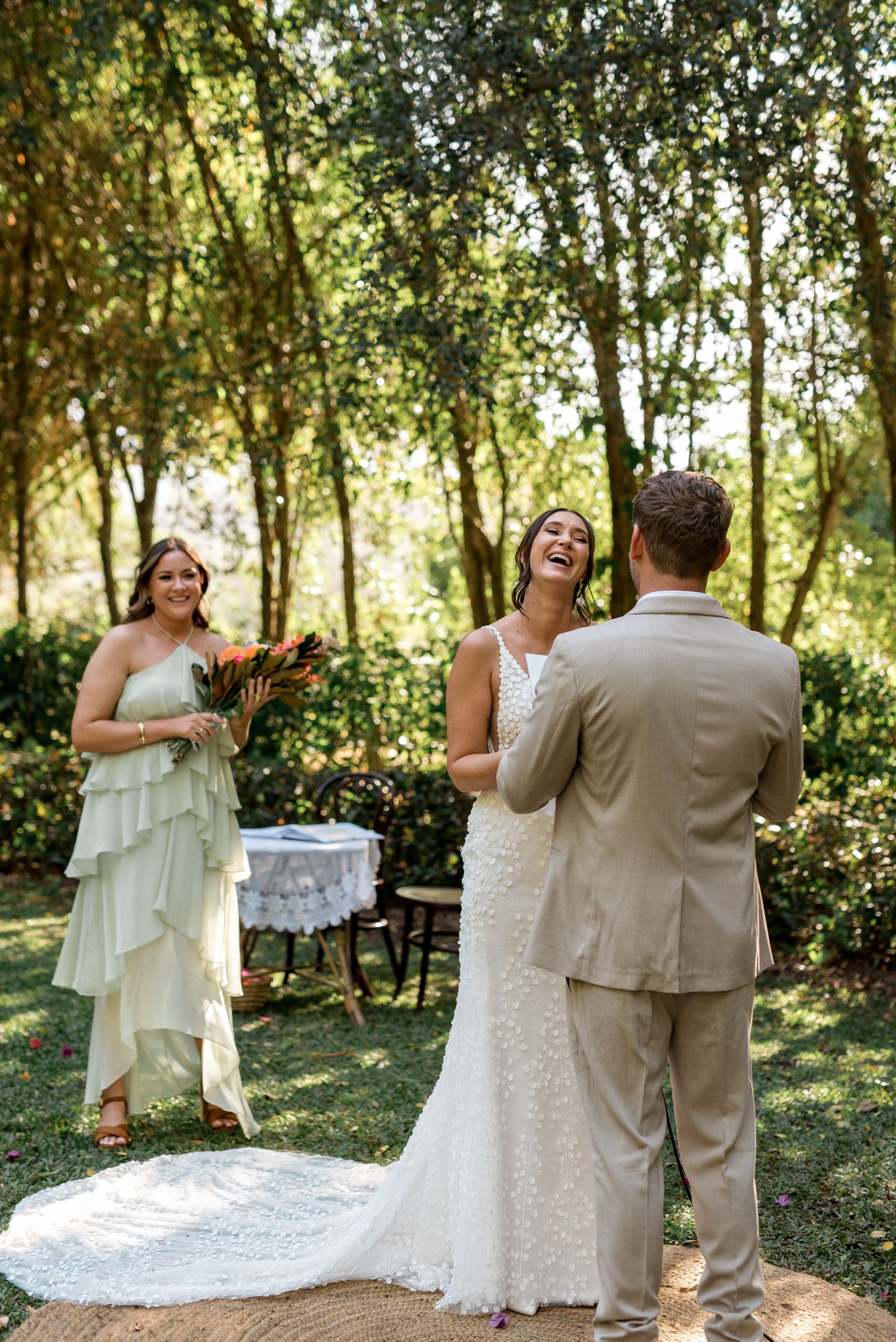 Bride laughs joyfully during outdoor garden wedding ceremony while bridesmaid smiles in sage green dress.
