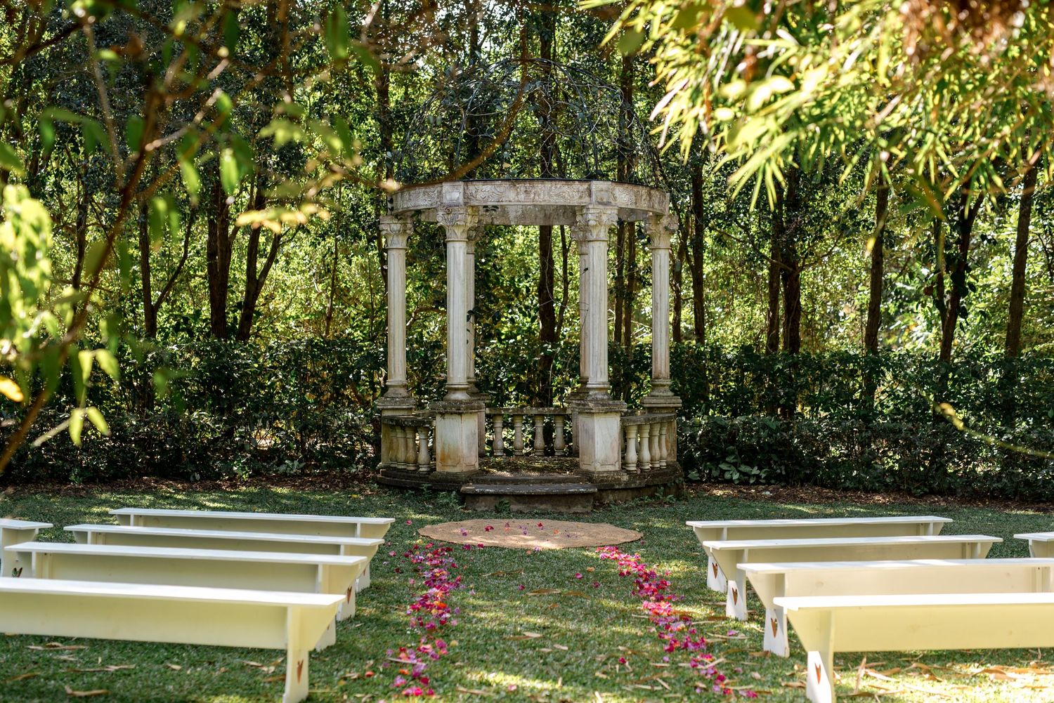 Elegant outdoor wedding ceremony setup with white benches, floral petal aisle, and stone gazebo.