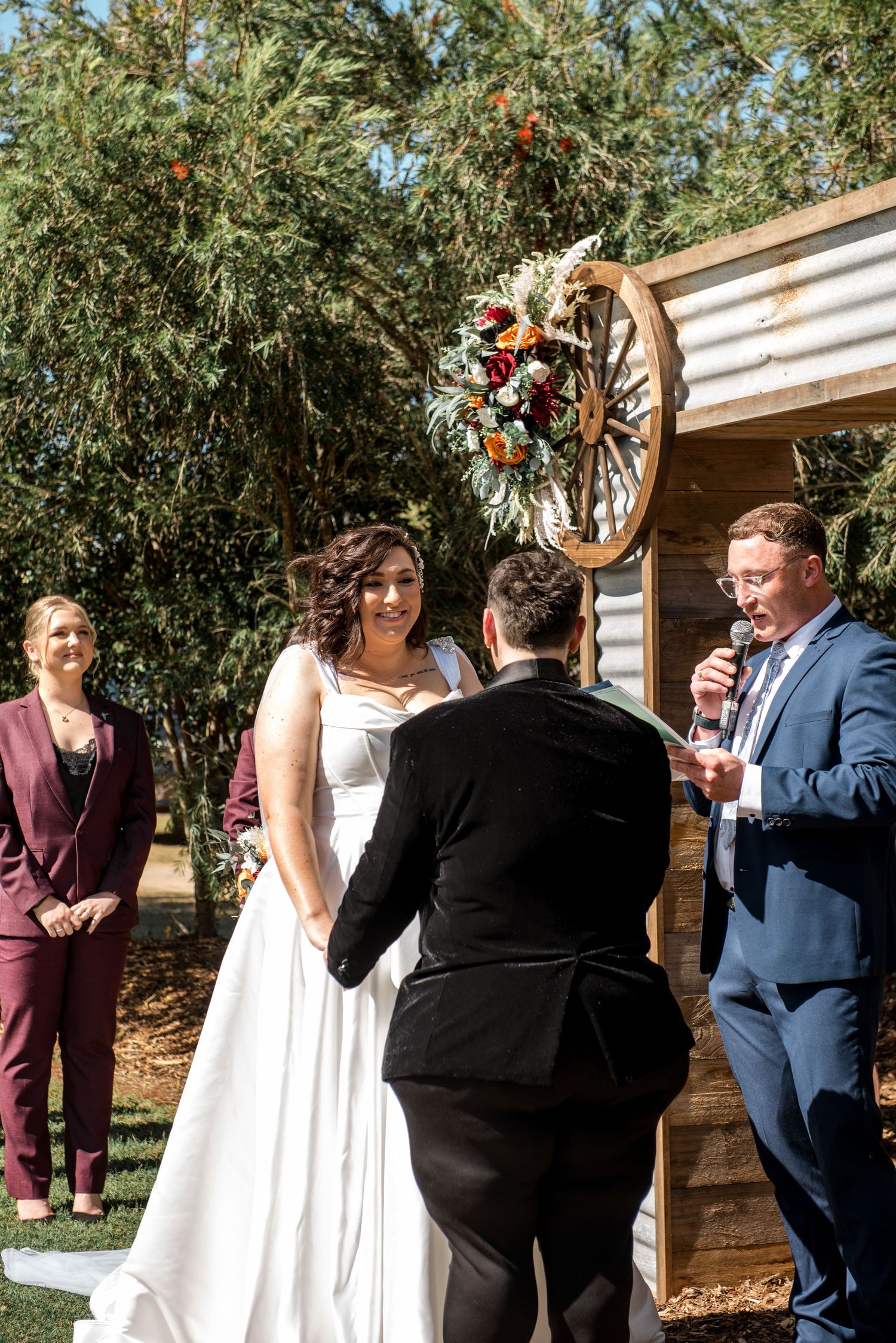 Bride and groom exchange vows at rustic outdoor ceremony with wagon wheel floral arch and wooden backdrop.