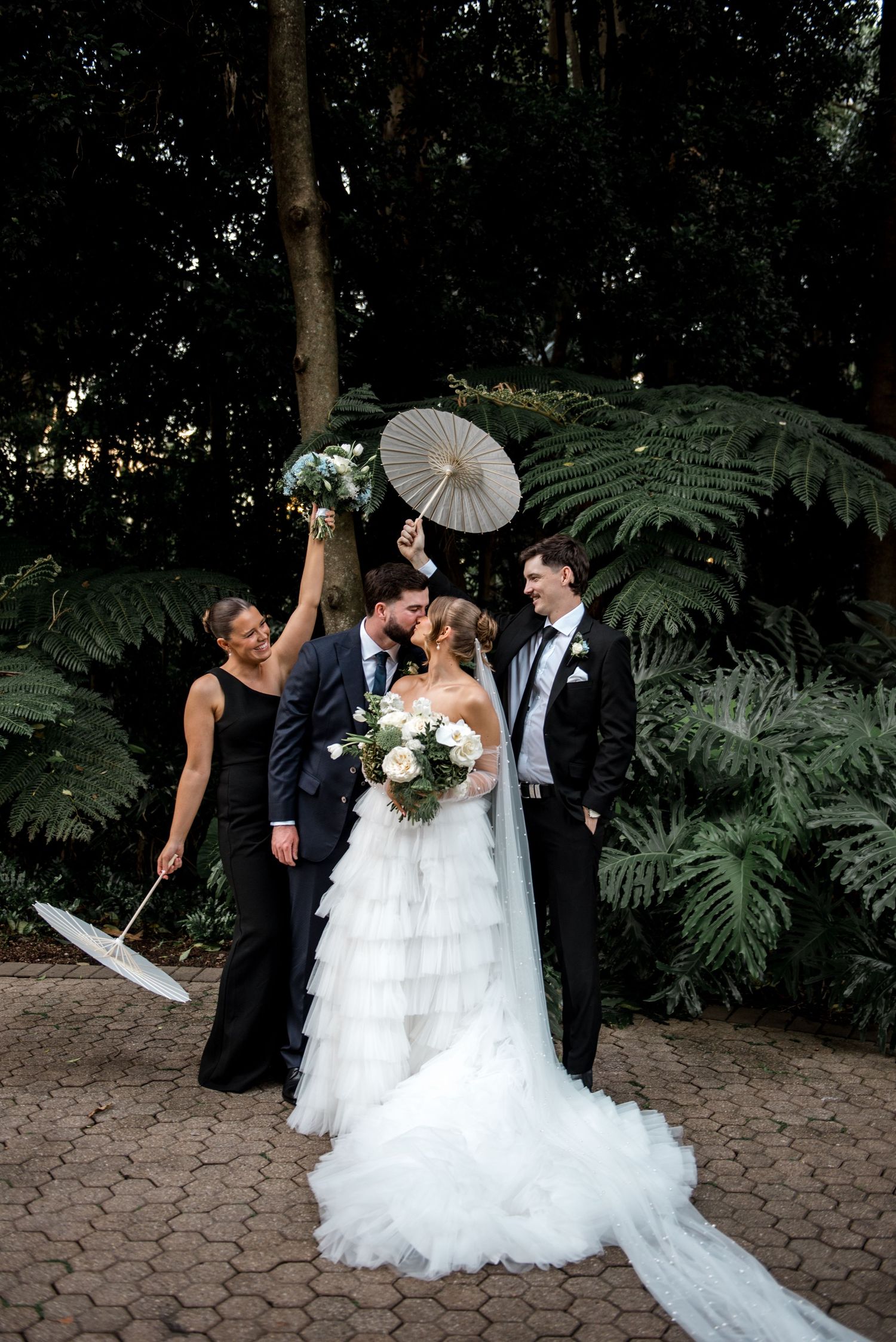 Wedding party celebrates with parasols as bride and groom kiss in a lush garden ceremony setting.