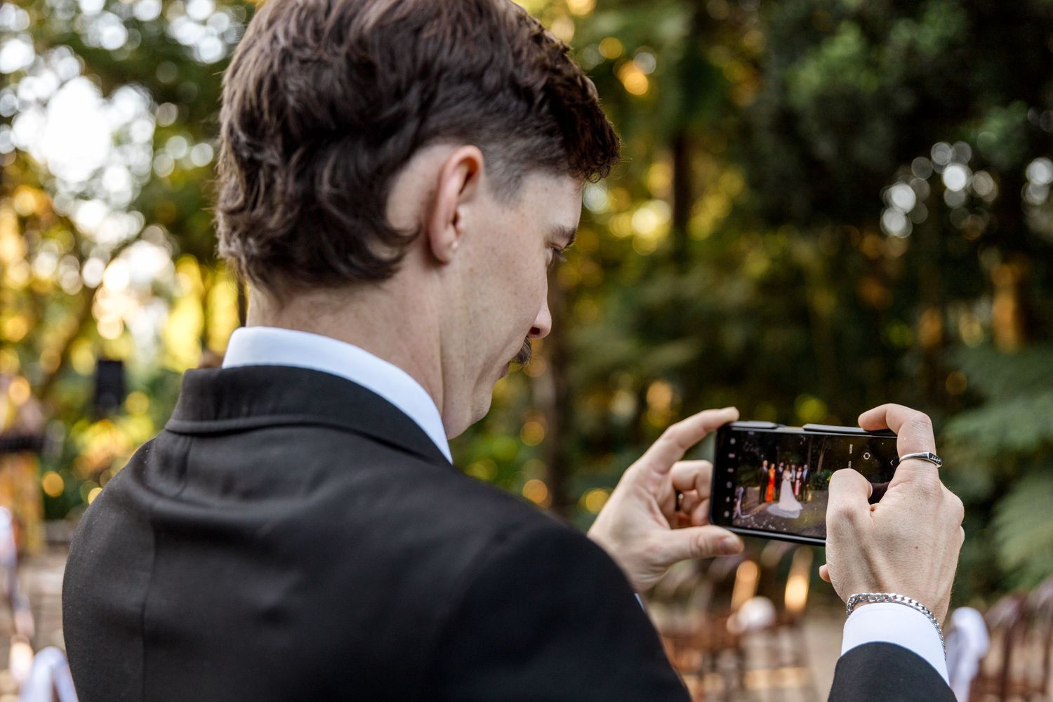 Guest photographs a wedding ceremony on his smartphone at an outdoor garden venue.
