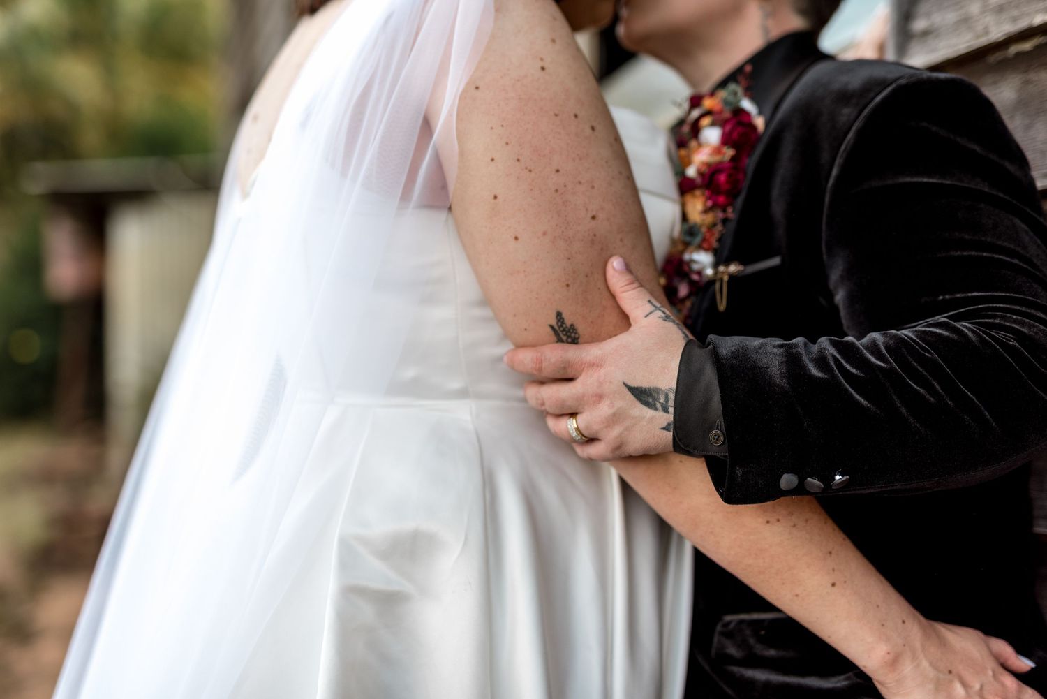 Close-up of groom in black velvet suit embracing bride in white wedding dress outdoors.