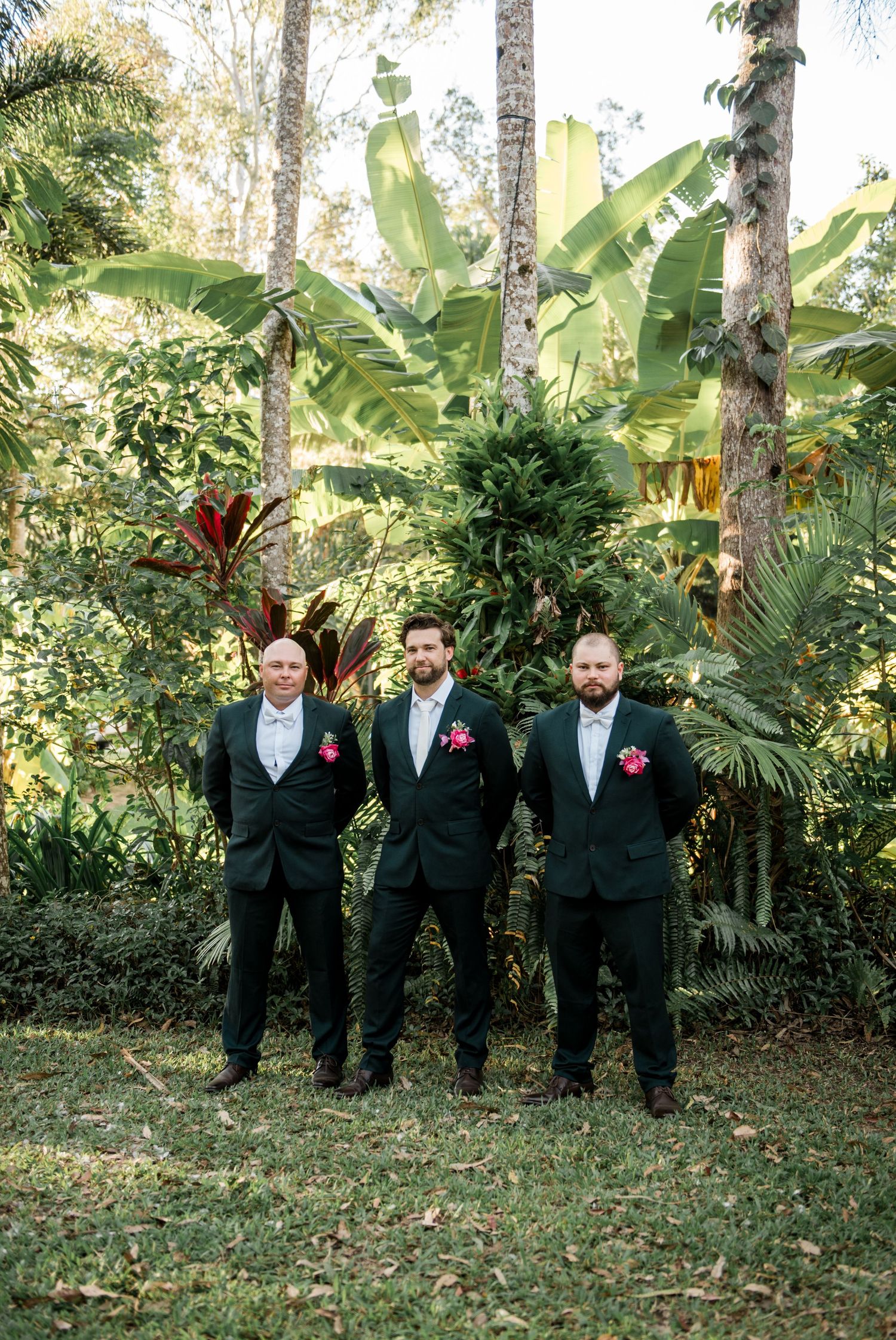 Three groomsmen in dark suits with pink boutonnieres pose before lush tropical foliage.