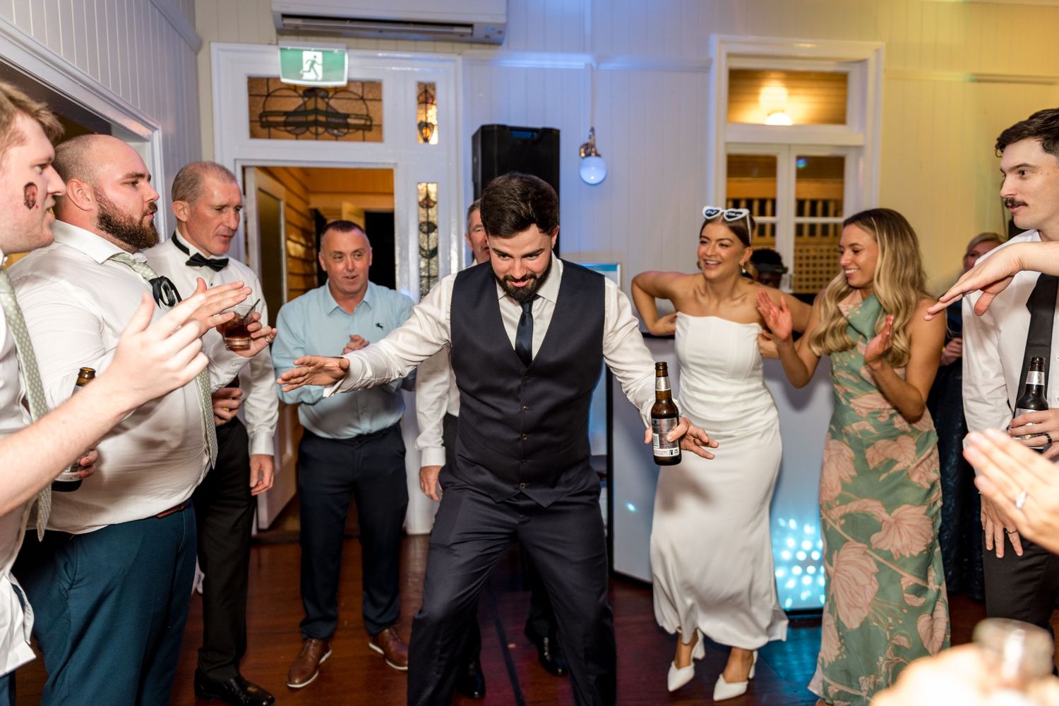 Groom in black vest dancing with beer bottle surrounded by cheering wedding guests.