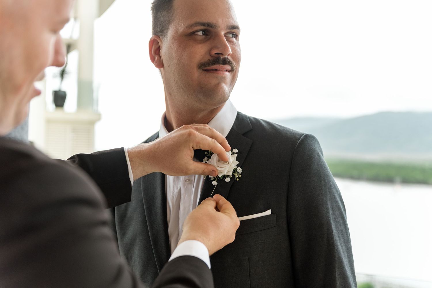 Best man pinning a white floral boutonniere on groom's black suit lapel outdoors.