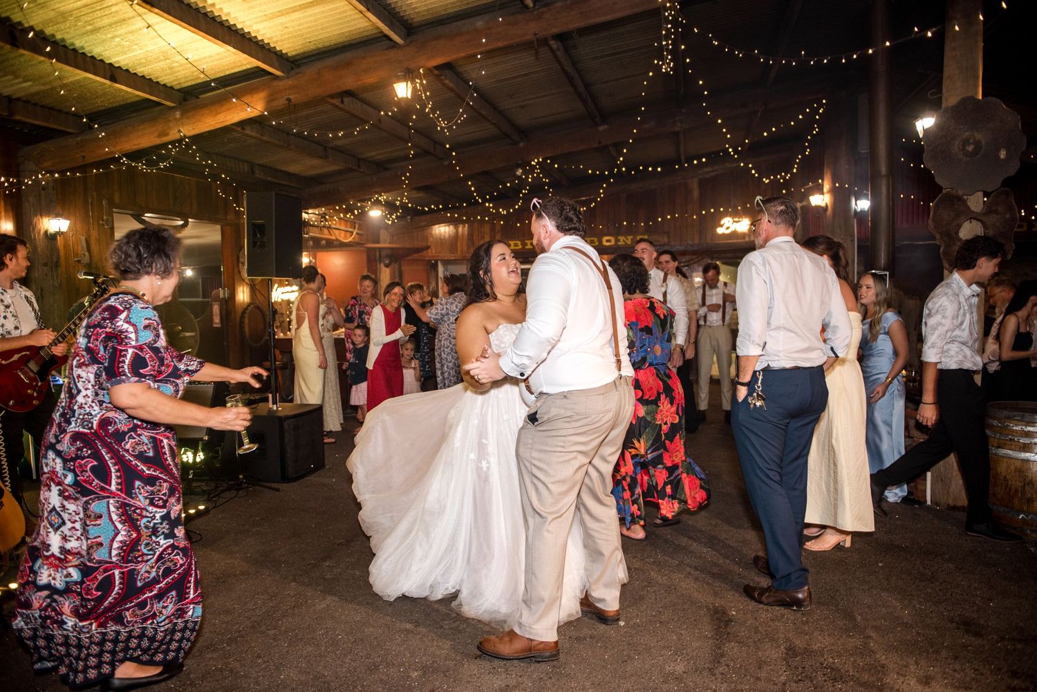 Bride and groom dance together at rustic barn wedding reception surrounded by guests.