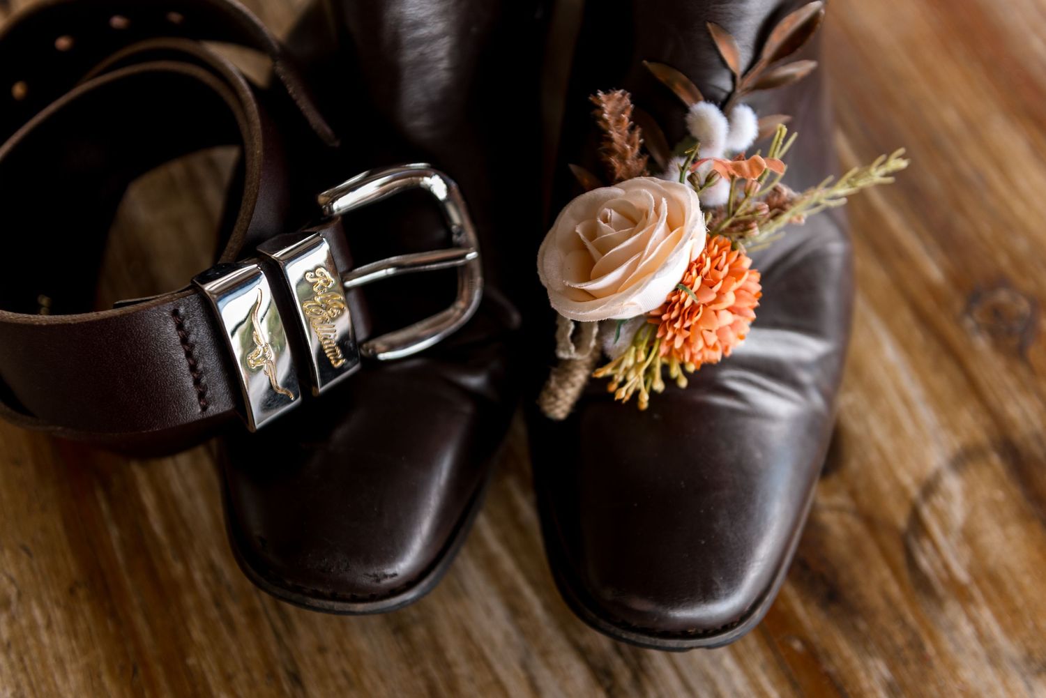 Brown leather cowboy boots with a floral boutonniere and silver belt buckle on a wooden surface.