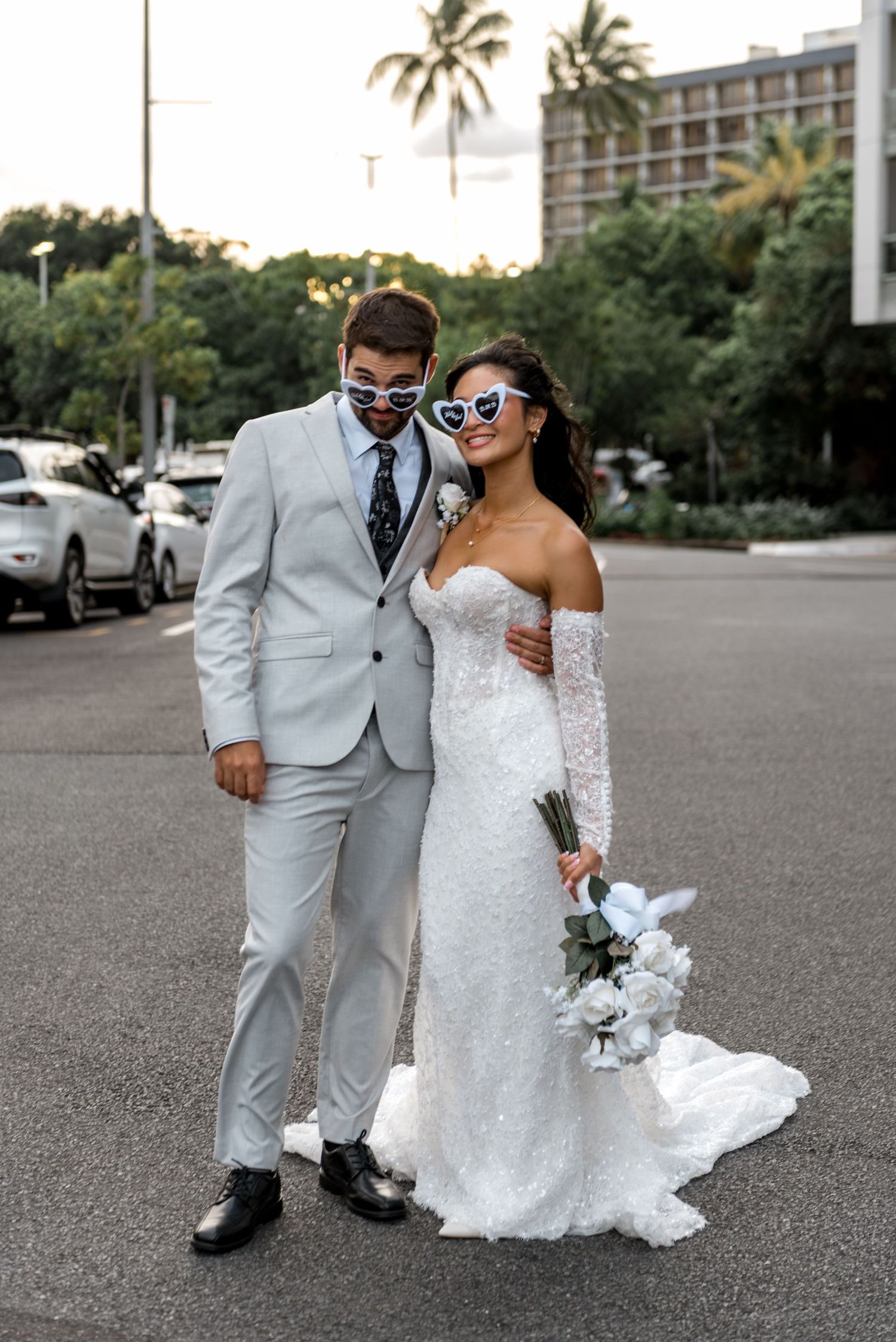 Bride and groom in light gray suit wearing heart-shaped sunglasses posing together on street with tropical background.