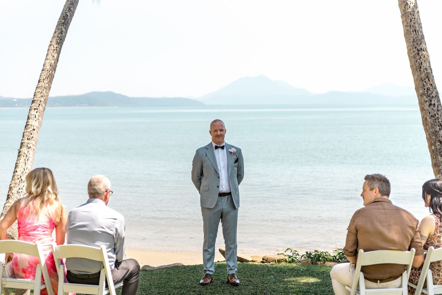 Groom in gray suit stands at beachside wedding altar with palm trees and ocean views.