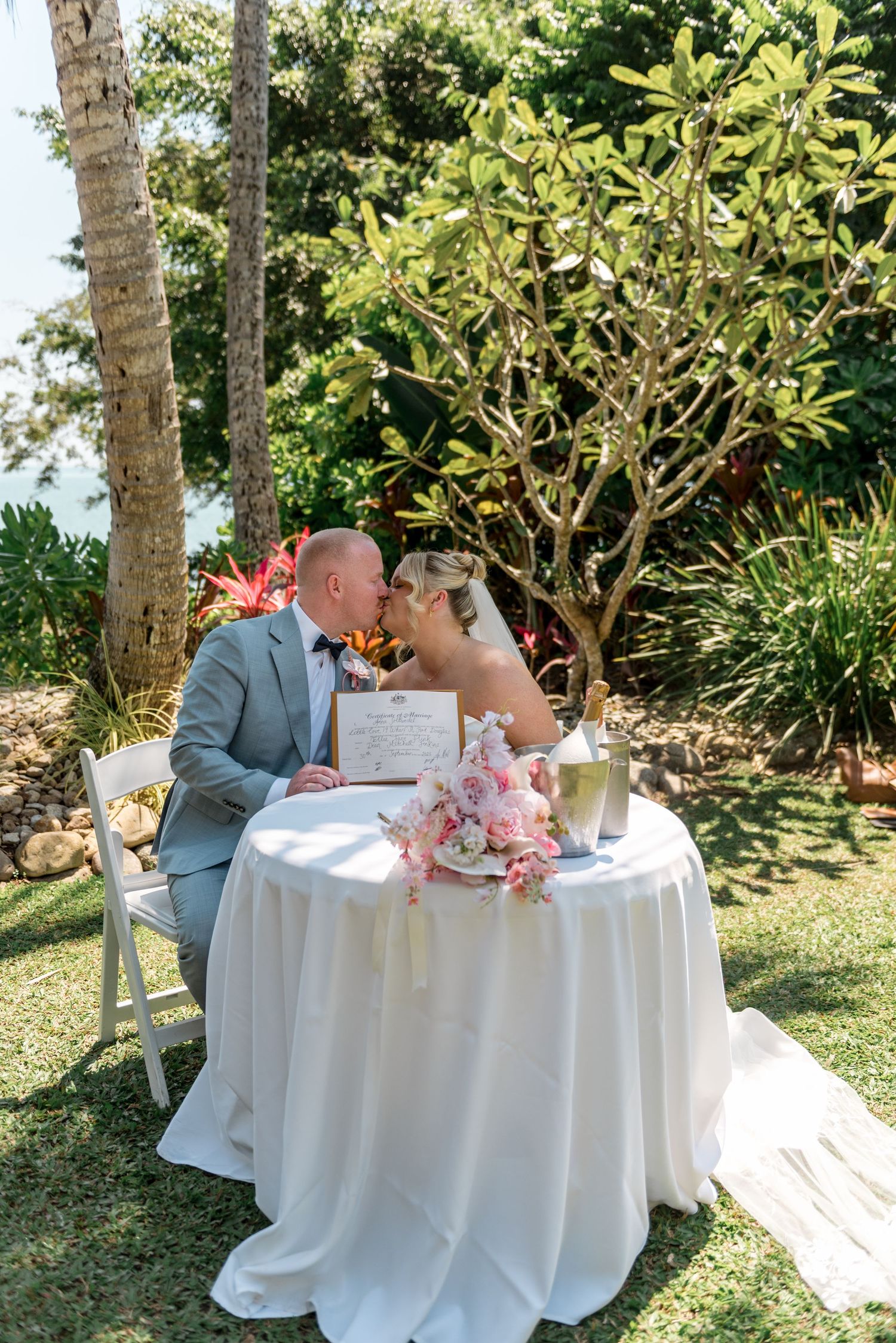 Newlywed couple sharing a kiss at signing table with pink bouquet and marriage certificate in tropical garden.