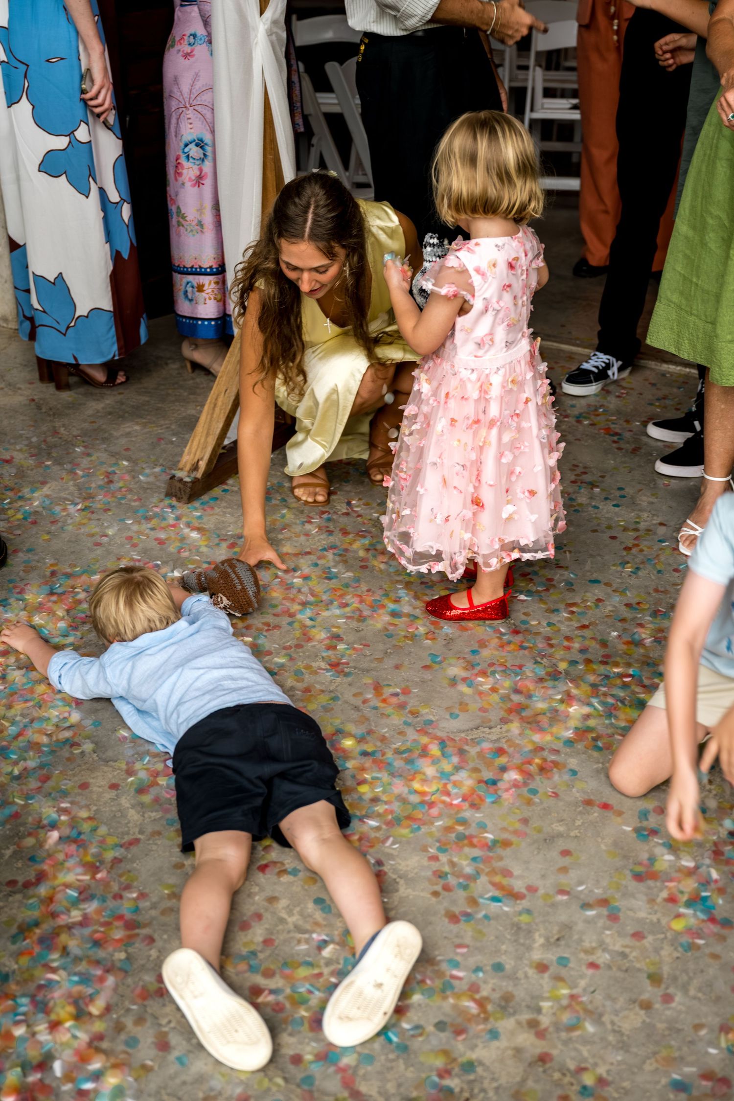 Woman in gold dress helps young children play with colorful confetti on ground at wedding.