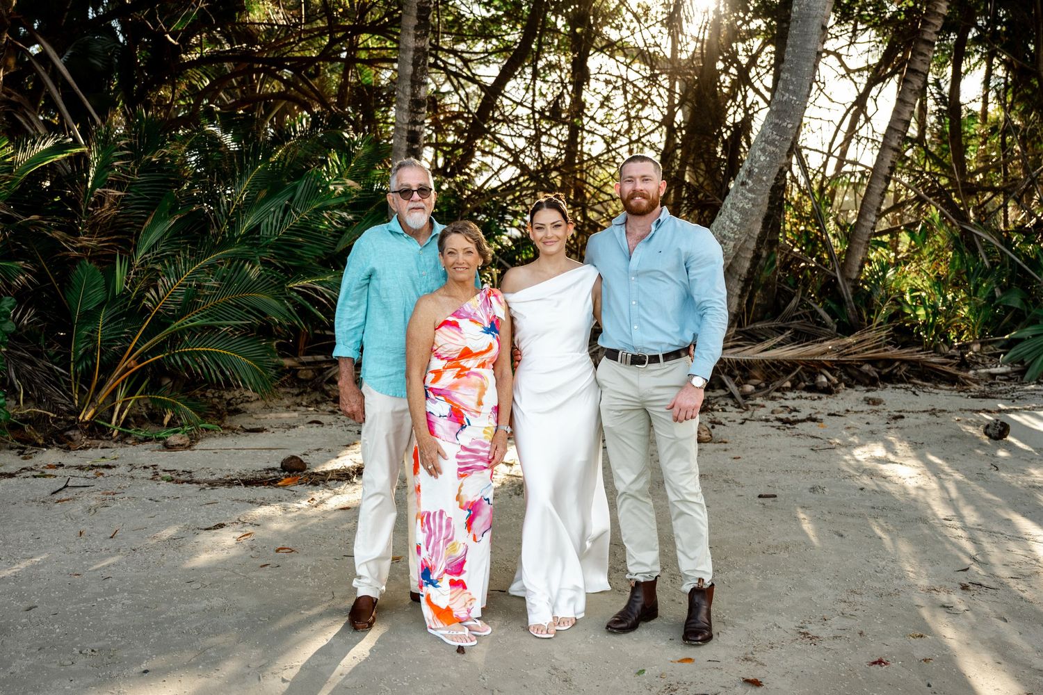 Family of four posing together on a sandy beach with lush tropical trees in background at Four Mile Beach, Port Douglas.