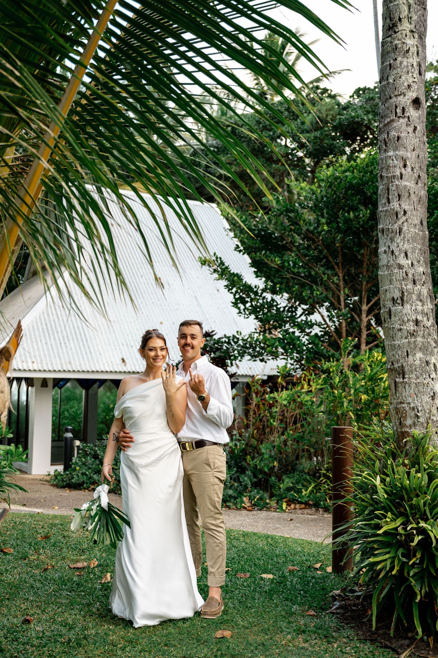 Smiling newlyweds showing wedding rings in a lush tropical garden beside a white chapel at Sheraton Grand Mirage Port Douglas.