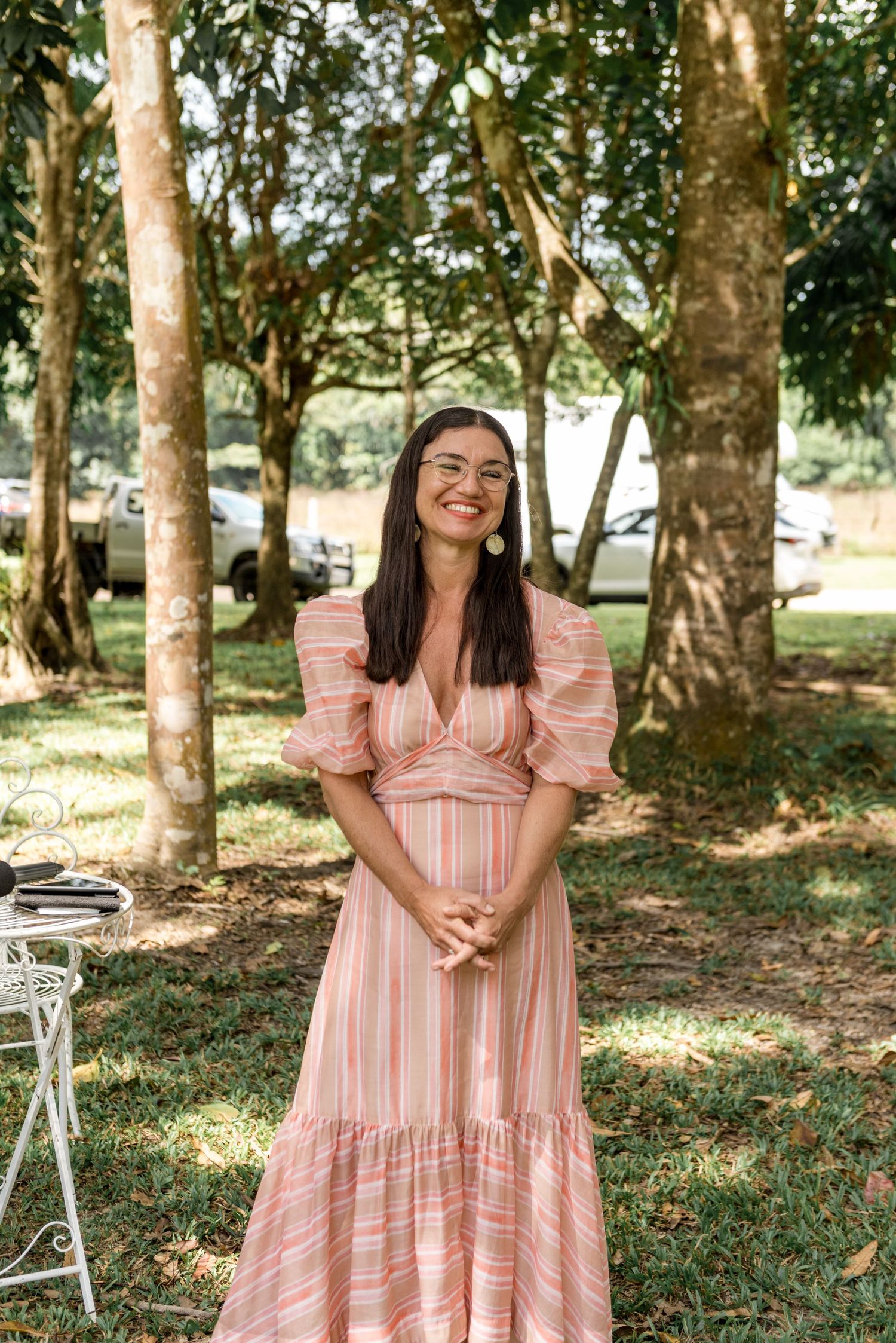 Smiling woman in striped pink dress stands outdoors among trees at a wedding celebration.