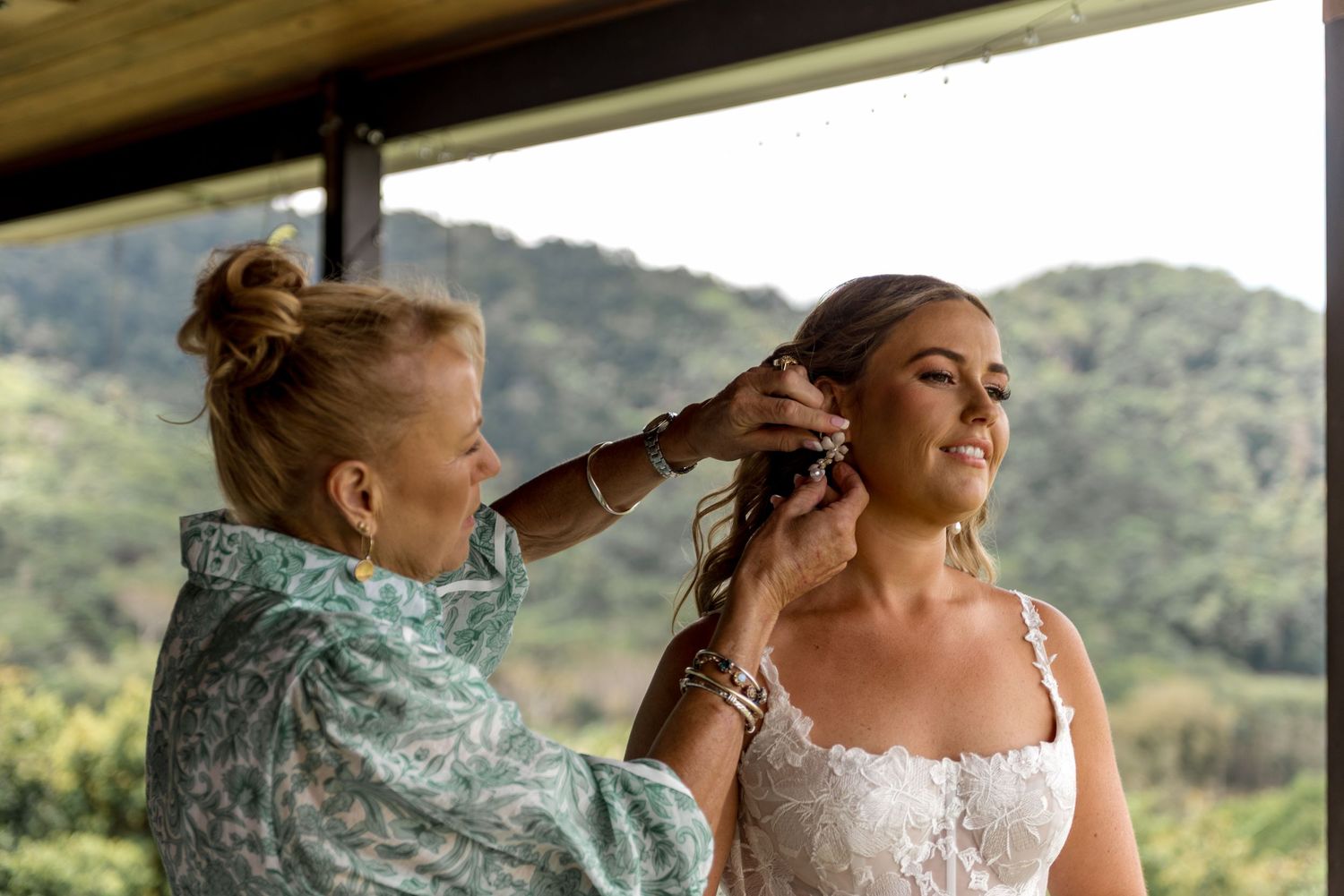 Hairstylist pinning bride's updo while bride smiles on a porch with green mountain scenery behind.