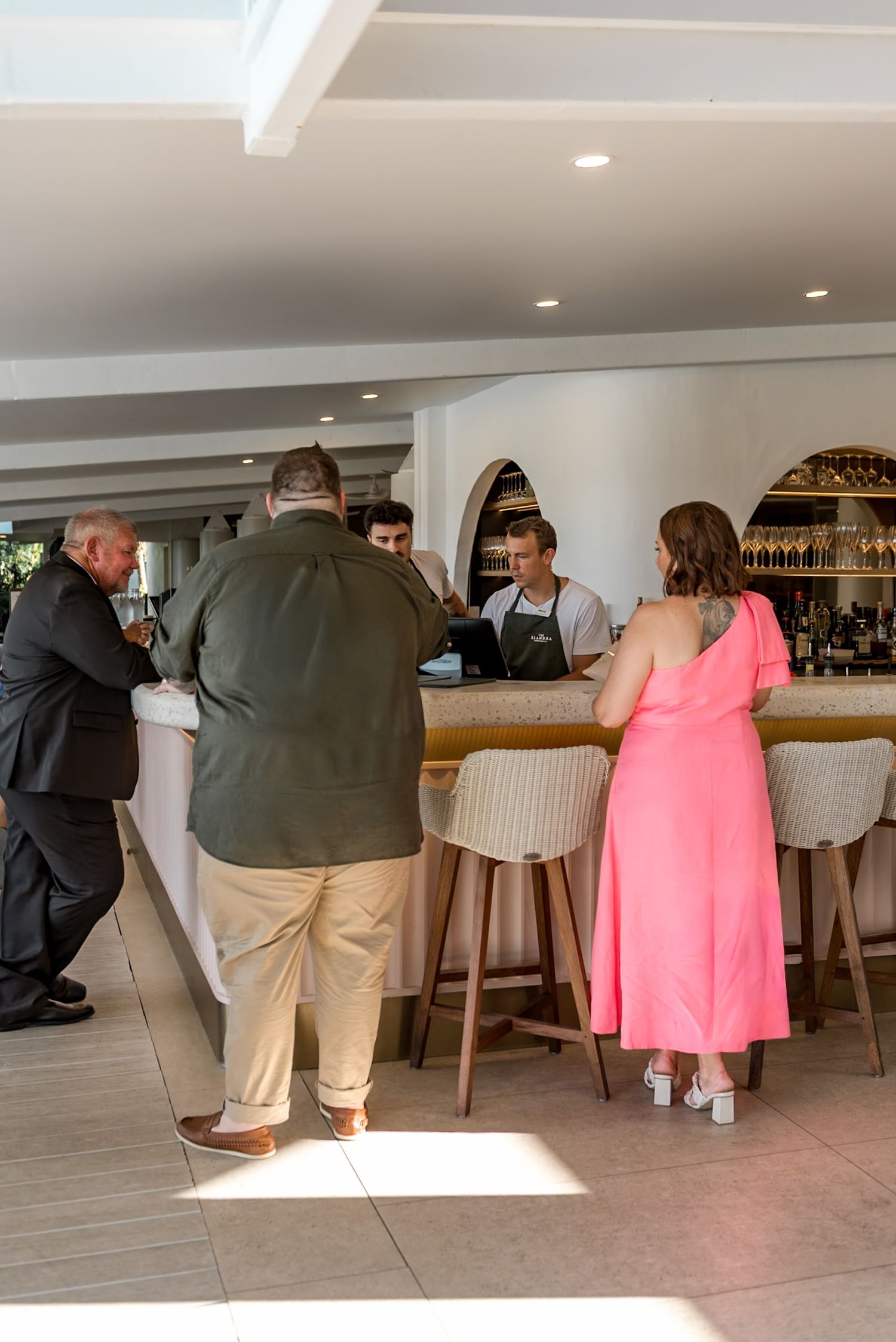 Wedding guests order drinks at elegant white bar with wicker stools at venue.