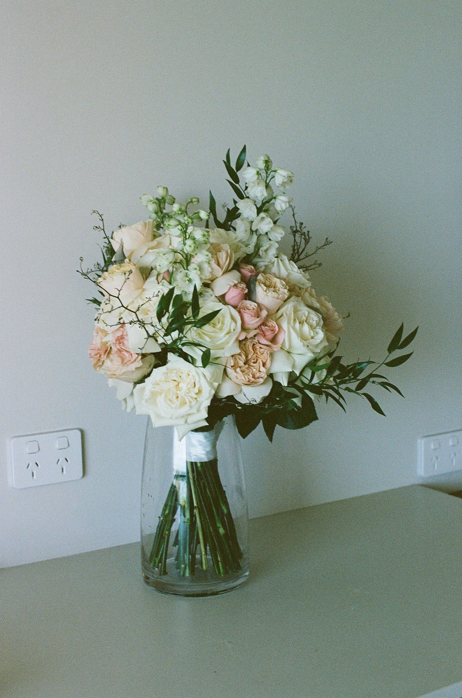 Romantic bridal bouquet of white and blush roses with greenery displayed in glass vase.