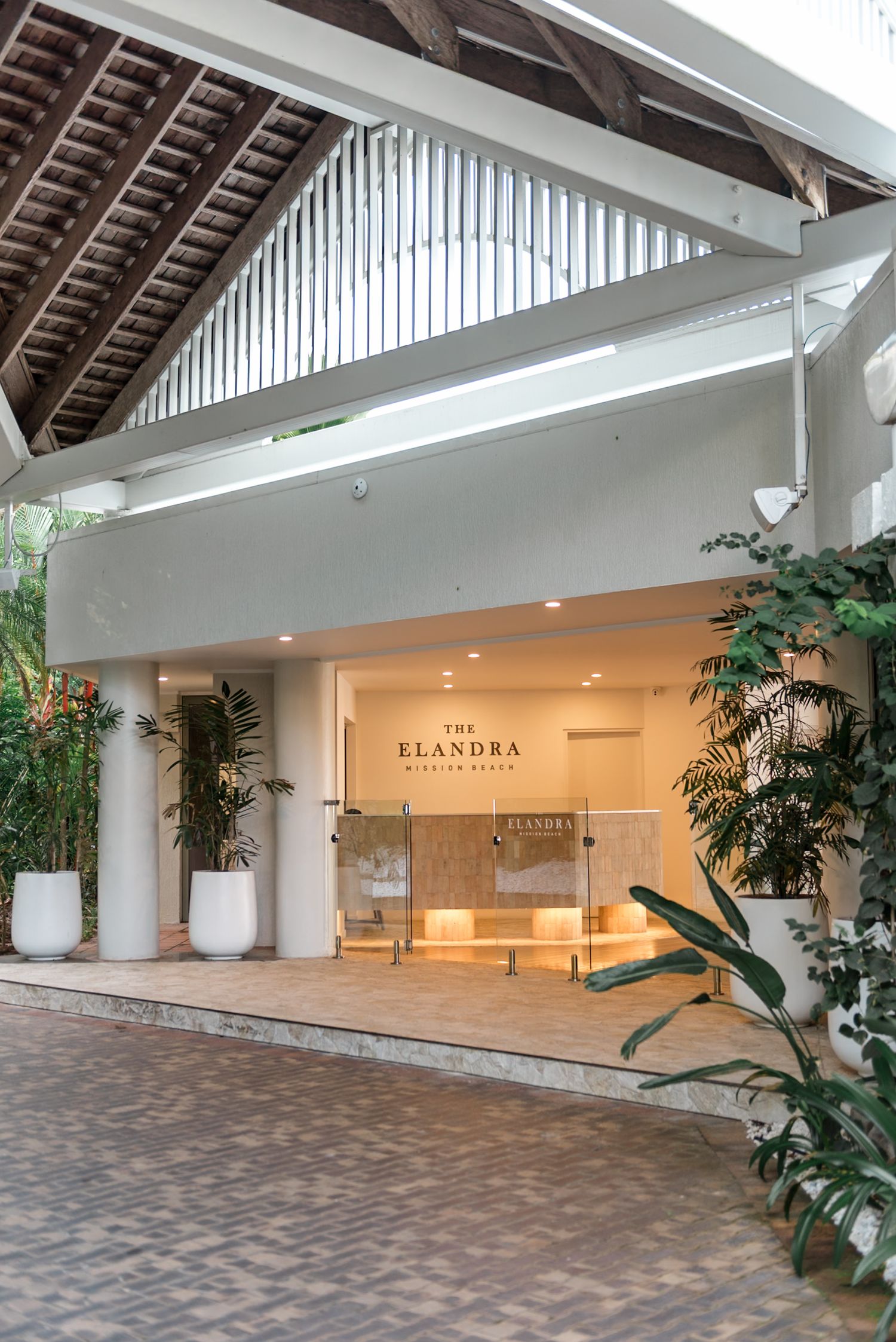 The Elandra Mission Beach hotel entrance with white walls, tropical plants, and warm interior lighting at dusk.