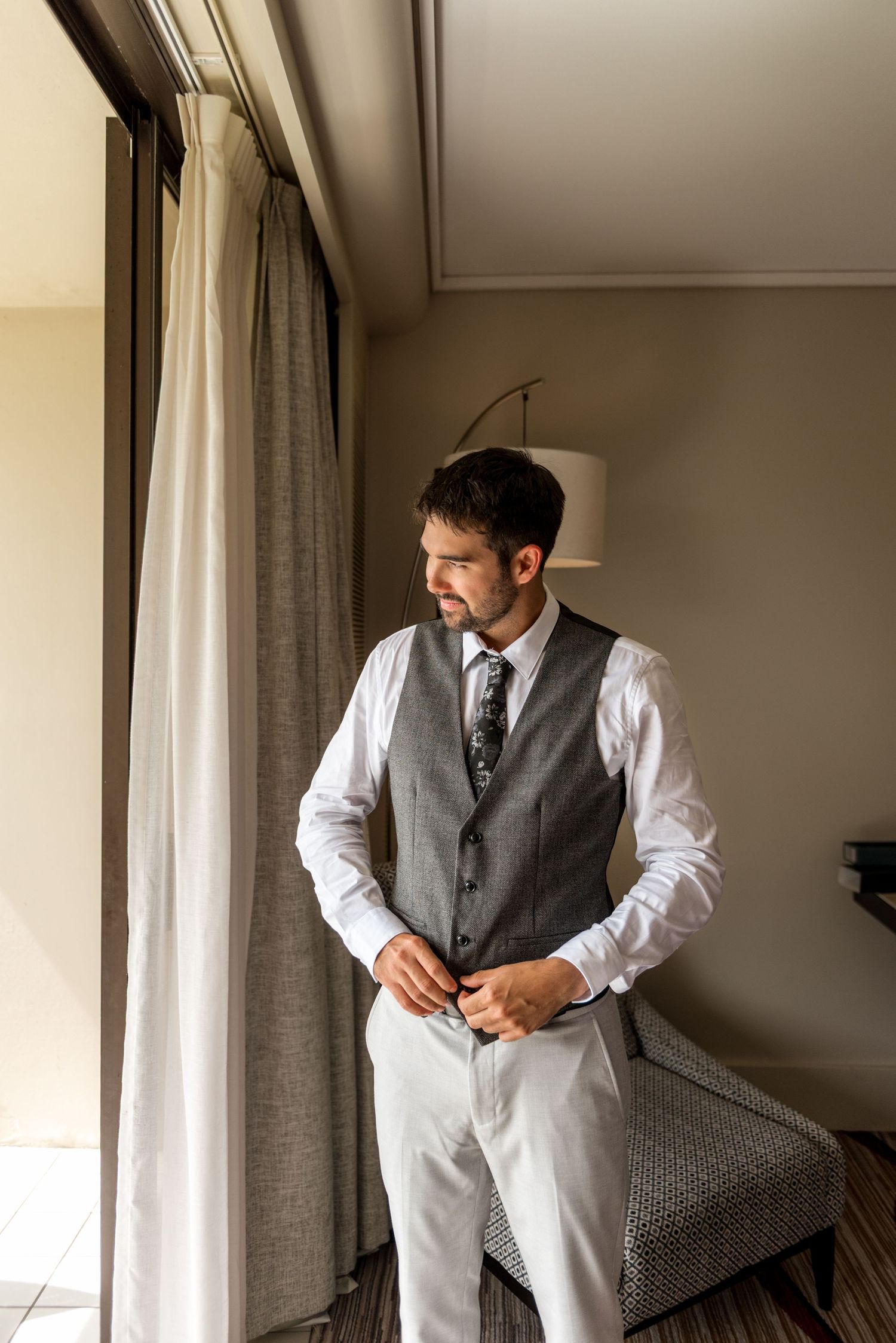 Handsome groom in light gray suit and floral tie gazing out a hotel window while getting dressed.