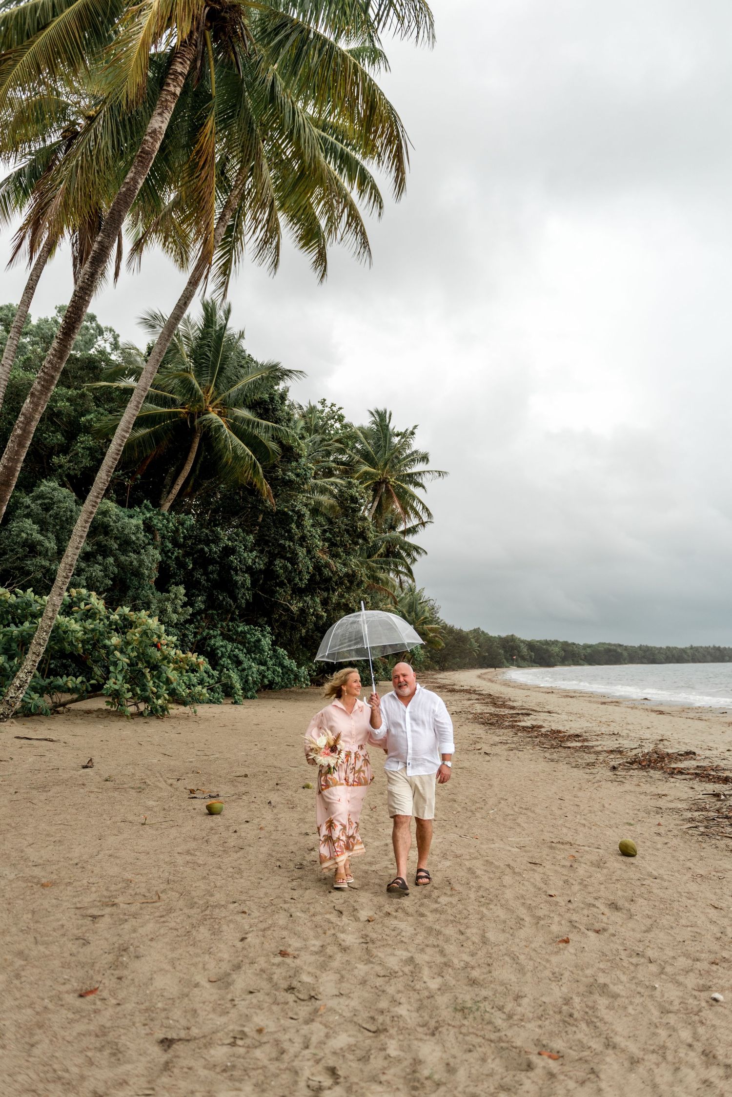 Couple strolling along a sandy tropical beach under an umbrella with lush palm trees nearby.