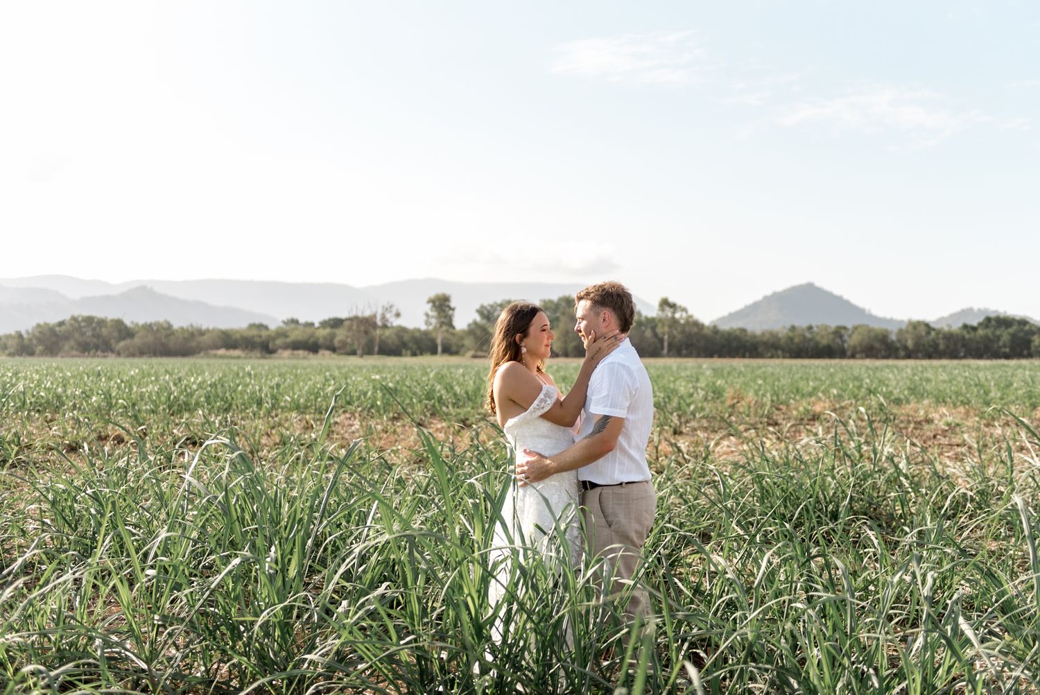 Couple embracing in a lush green field with mountains in the background on a sunny day.