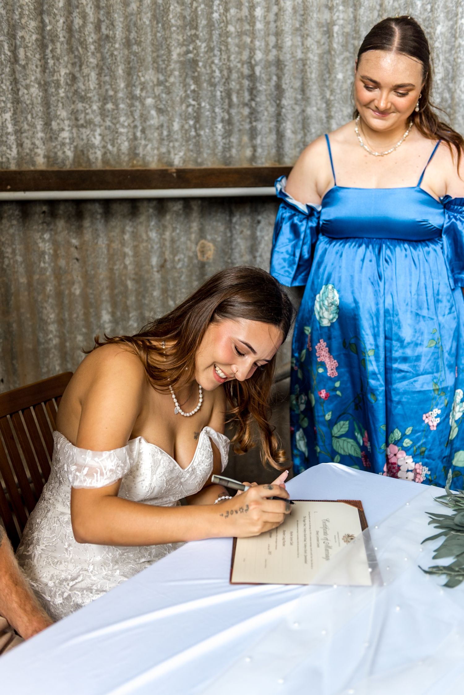Smiling bride signs marriage certificate while bridesmaid in blue dress watches.