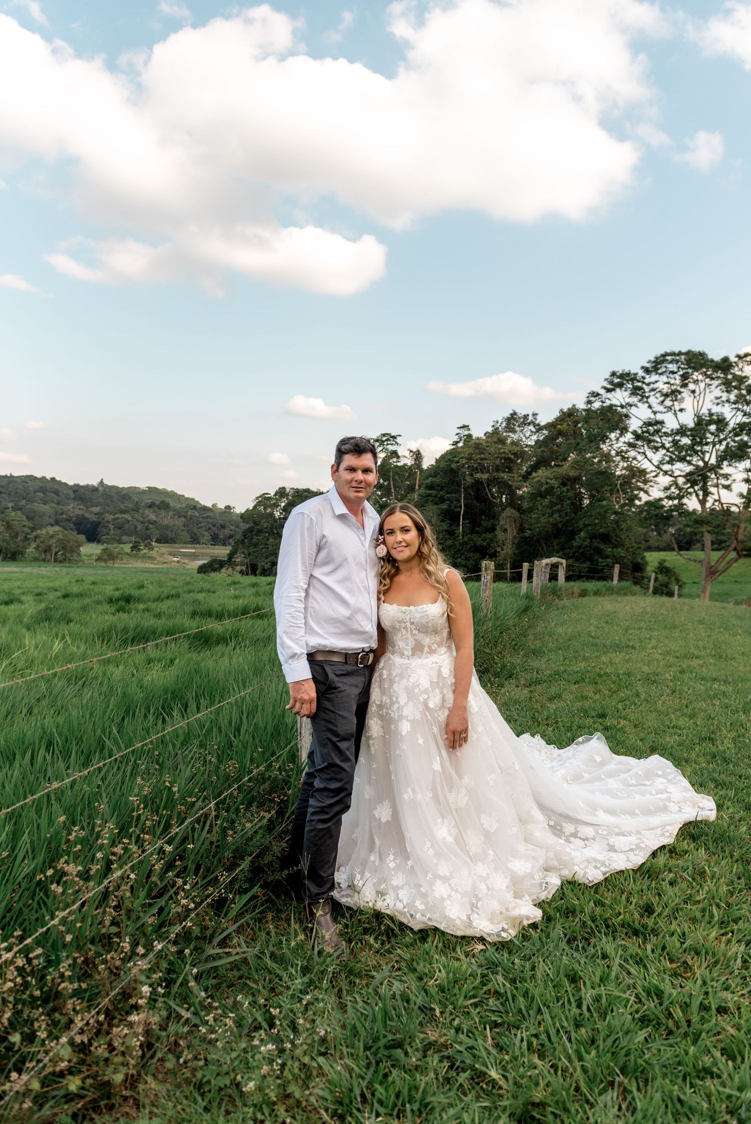 Bride and groom posing in a green field with dramatic clouds and tropical trees in background.